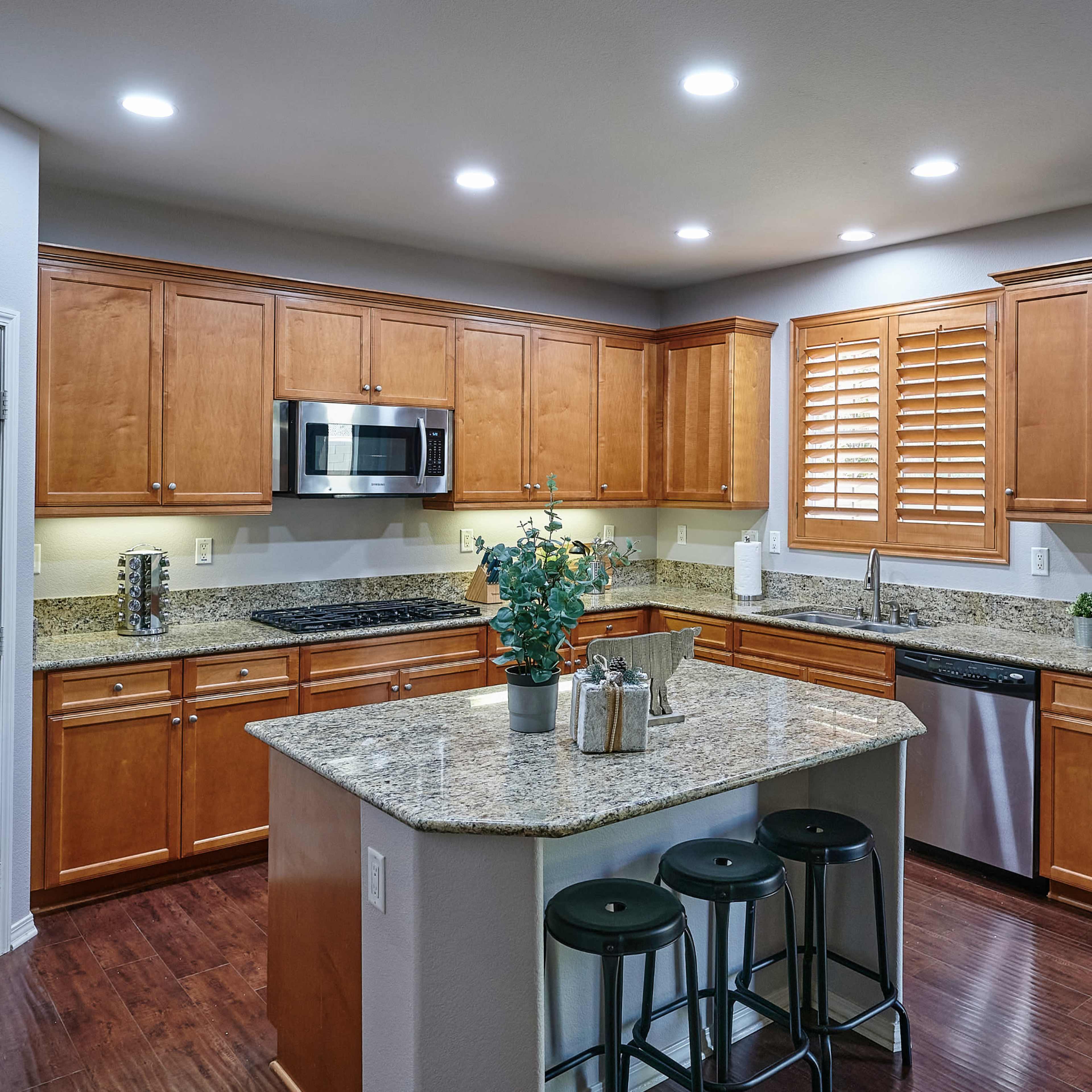 A modern kitchen features wooden cabinetry, granite countertops, and stainless steel appliances with a central island and bar stools.