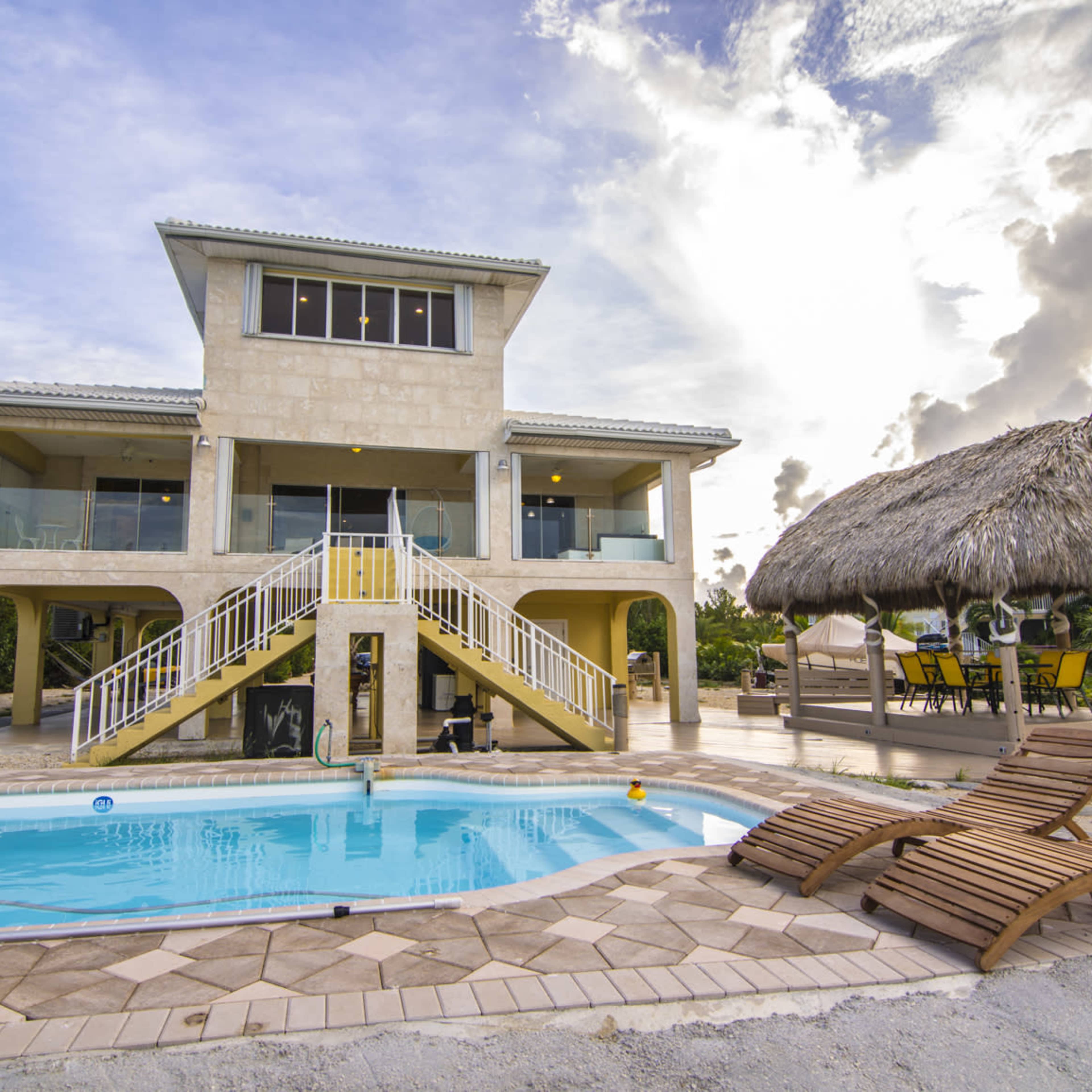 A modern two-story villa with a swimming pool is situated next to a thatched-roof gazebo in a scenic outdoor setting.