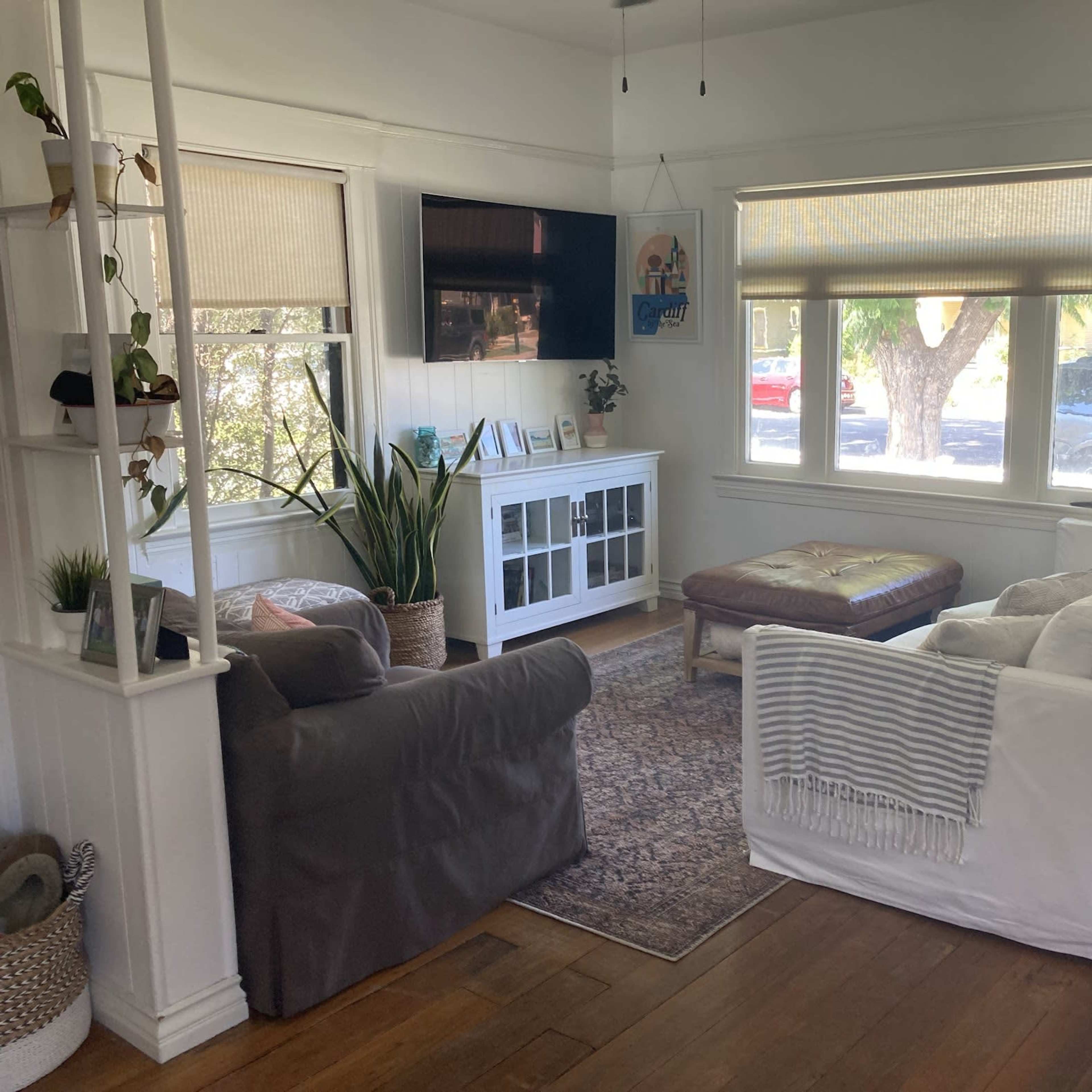 A living room with a grey armchair, a white couch, a coffee table, and a television mounted on the wall, all situated near large windows with blinds.