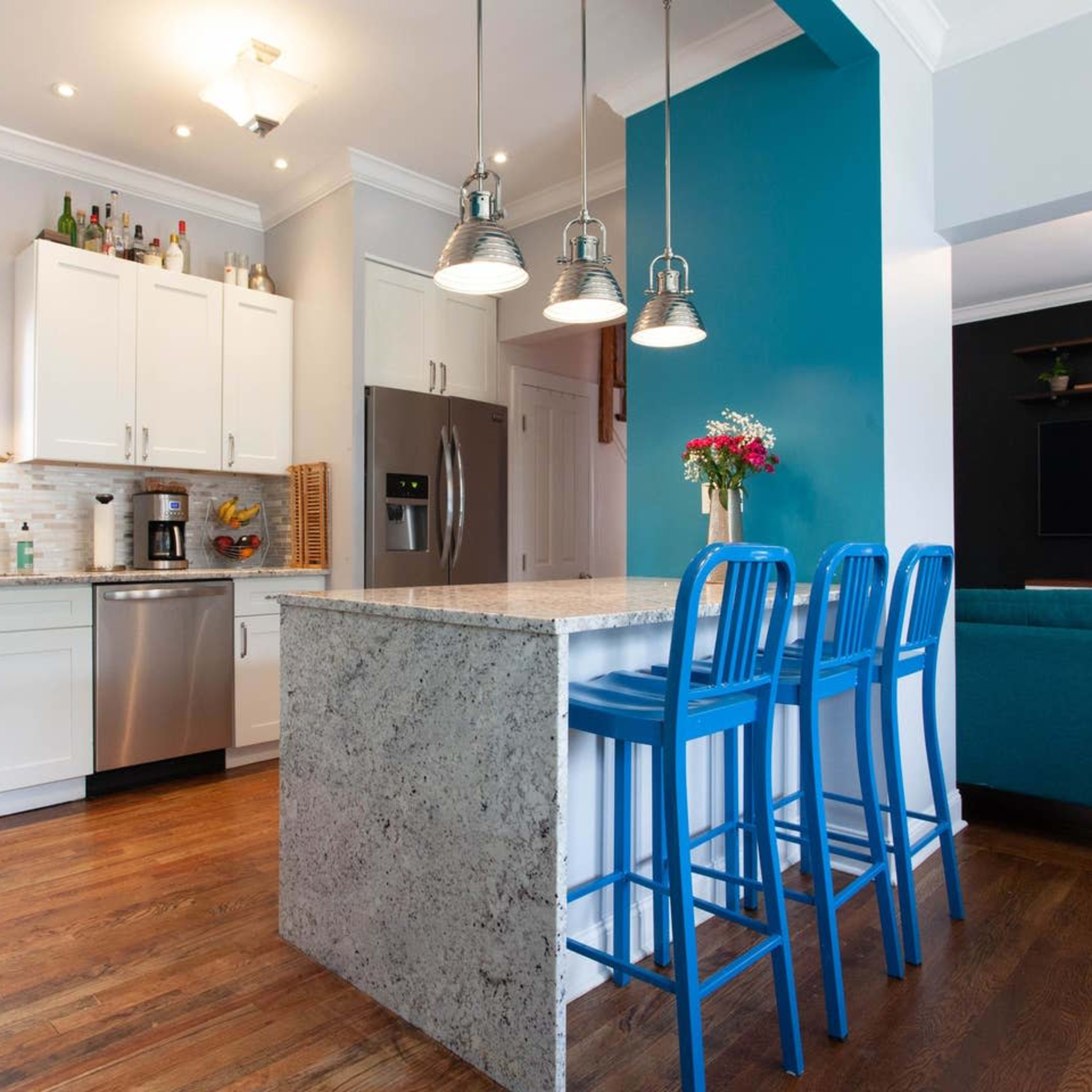 A modern kitchen features white cabinets, stainless steel appliances, and a blue accent wall with three blue bar stools at a stone island.