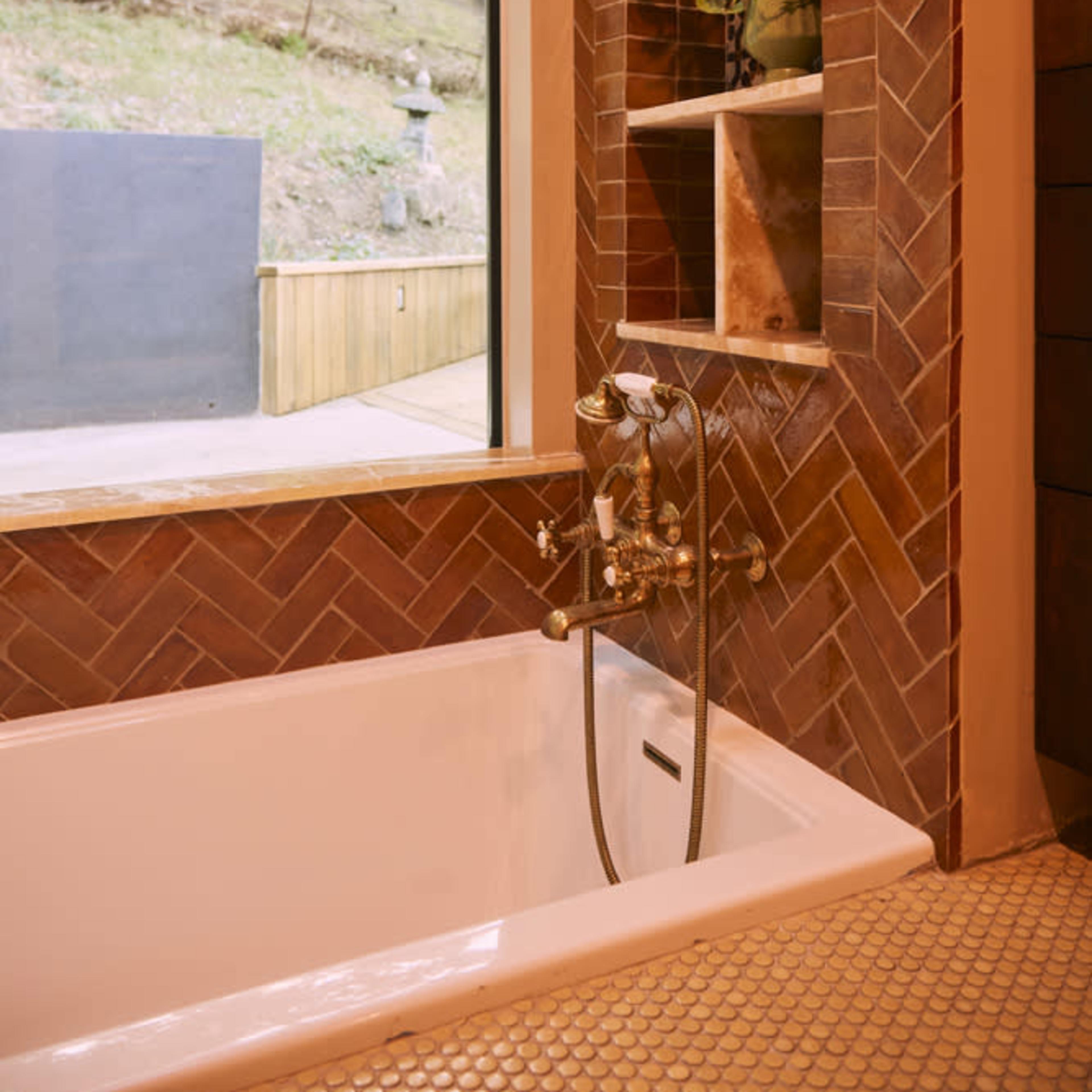 A modern bathtub with a herringbone-patterned backsplash is positioned beside a large window overlooking a green hillside.