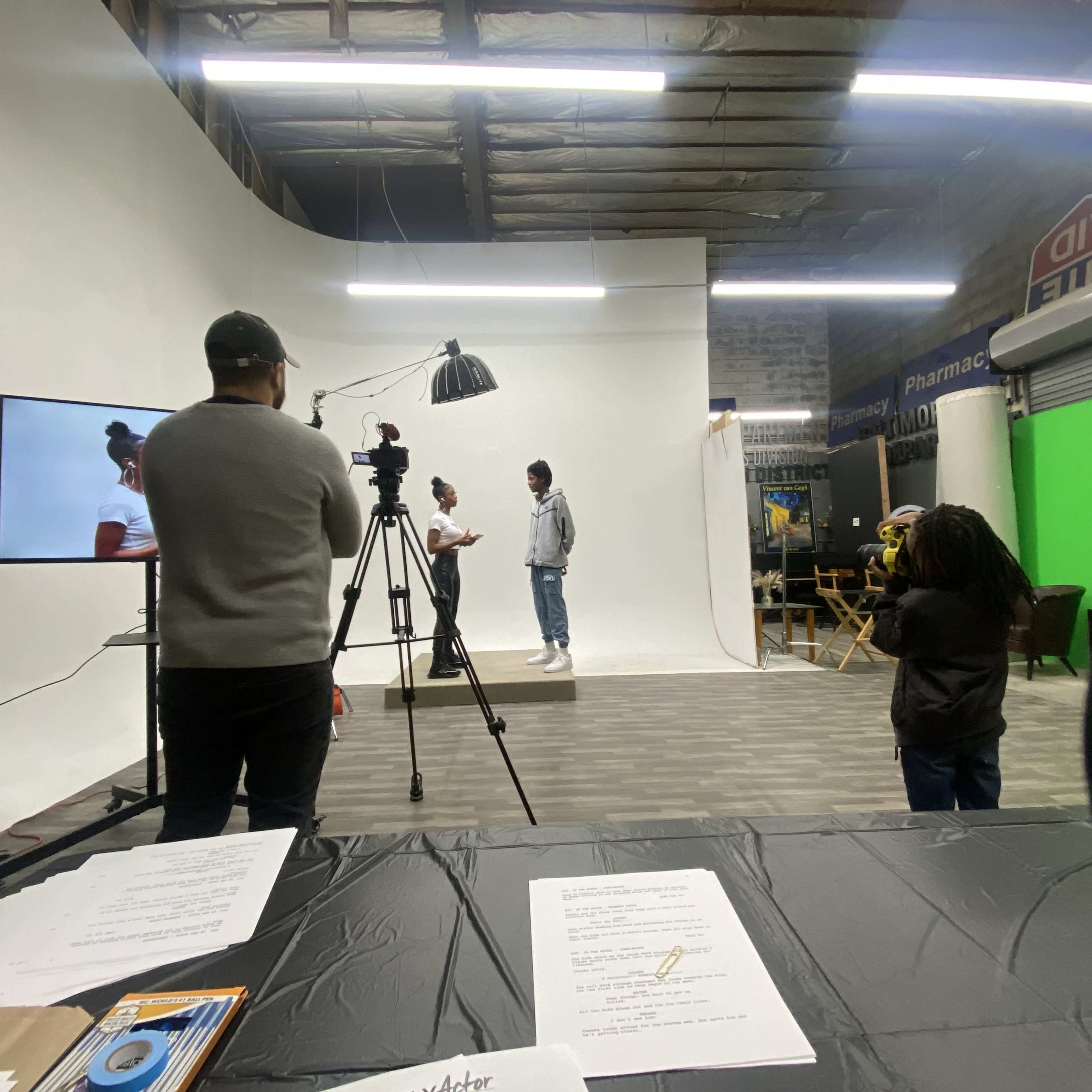A group of people is filming a scene in a studio with a white backdrop, while another person operates a camera.