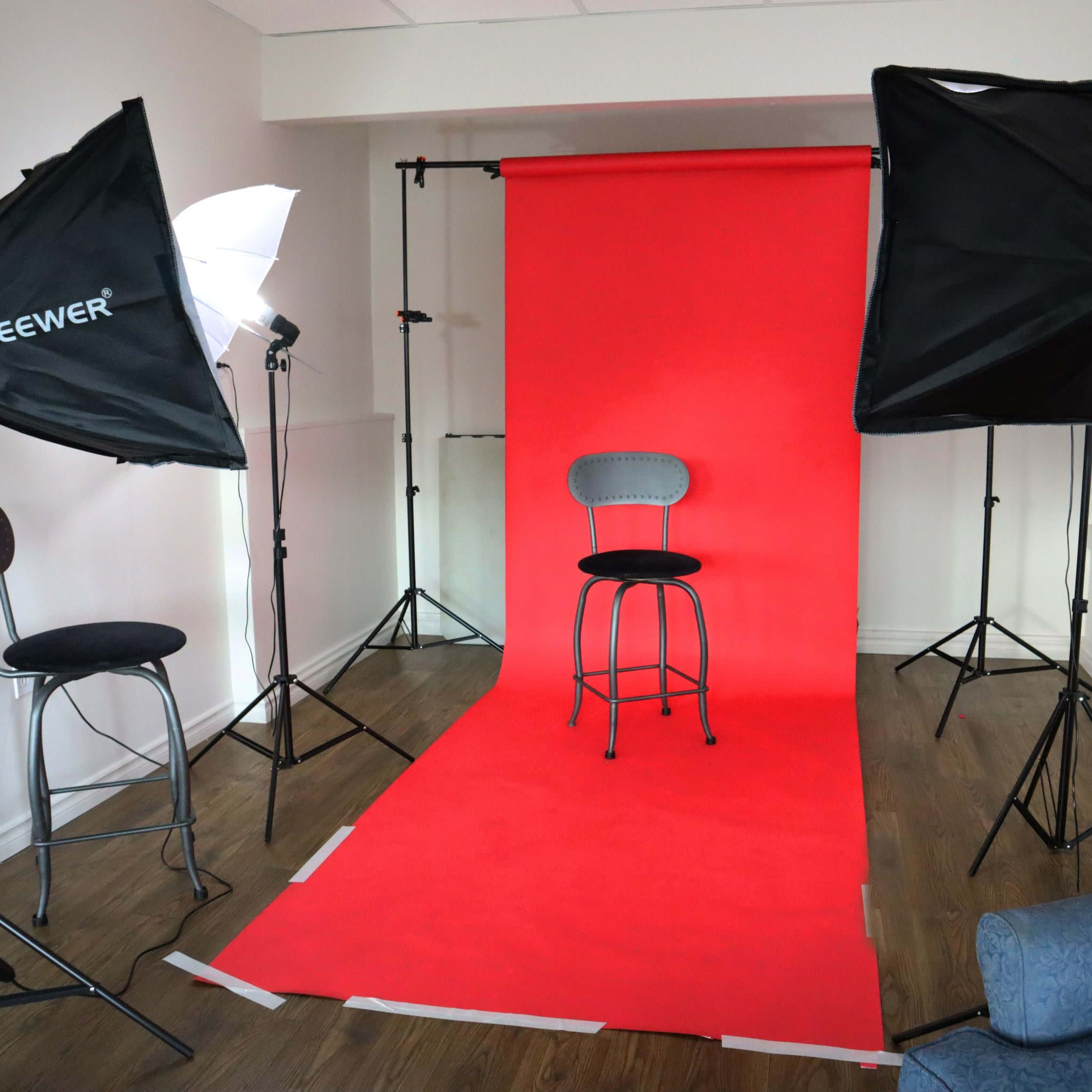 A photo studio set up with a red backdrop, three lighting setups, and a single black stool in the center.