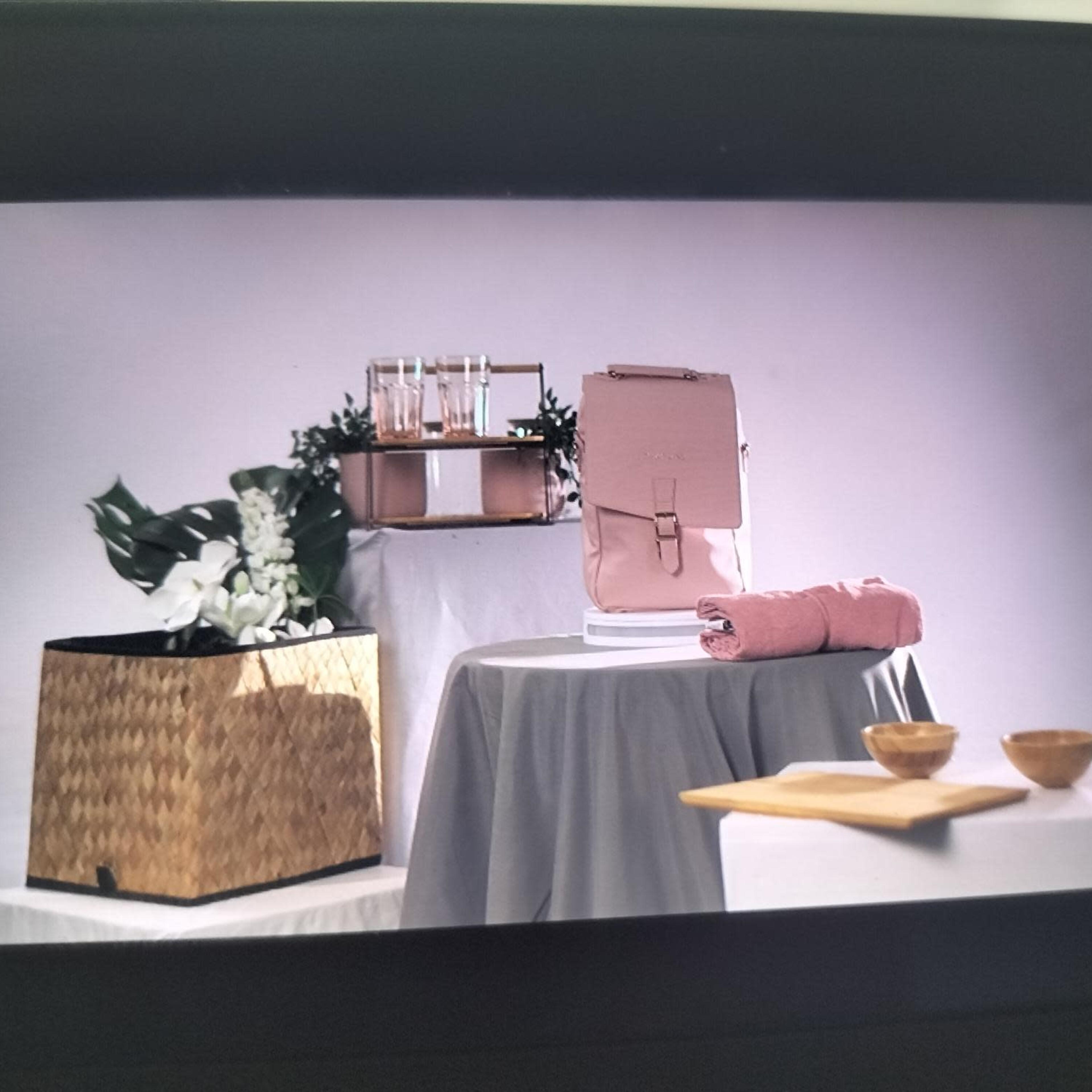 A pink backpack on a white display table next to a woven basket, a rolled towel, two wooden bowls, and glasses, all set against a plain backdrop.