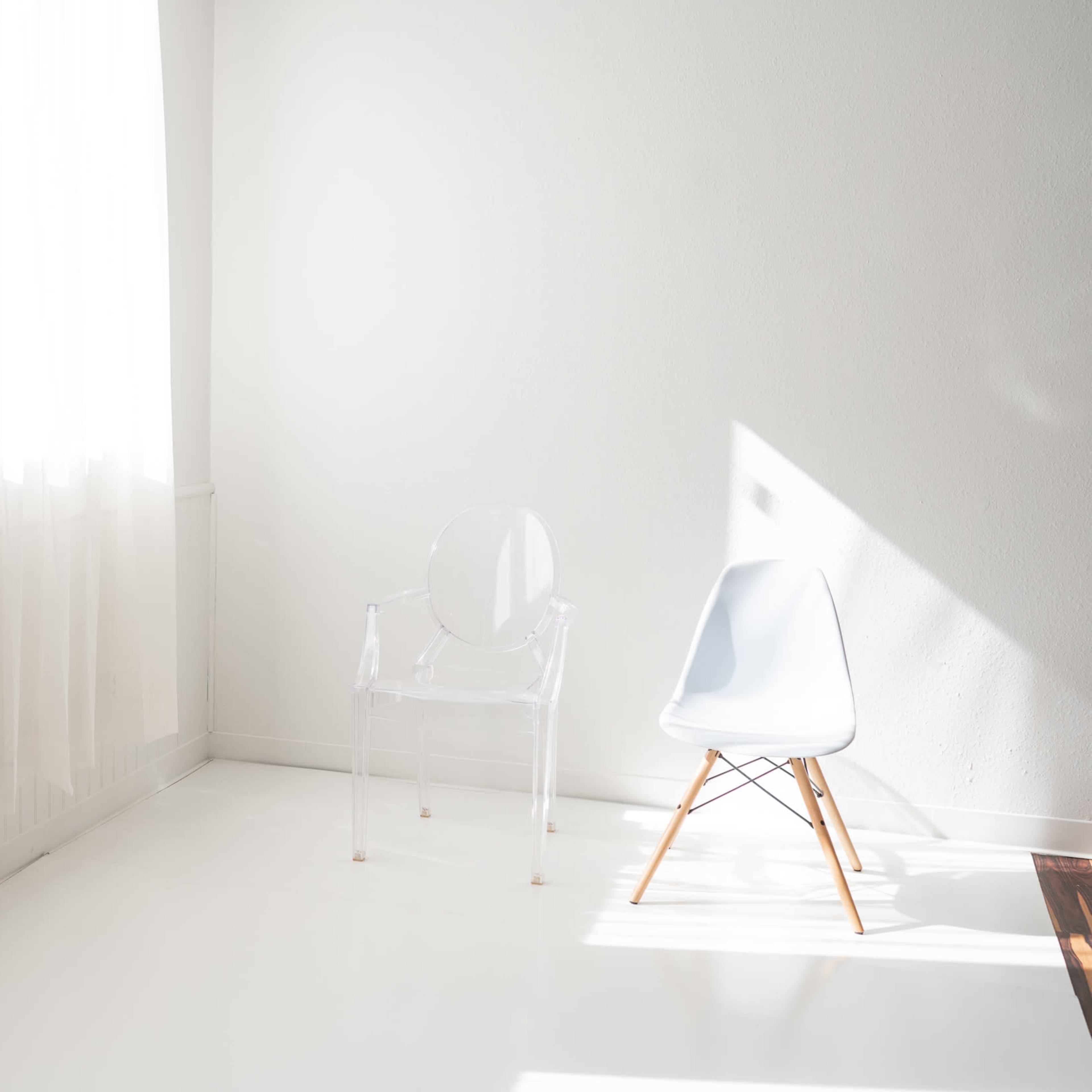 A bright, minimalist room features a clear acrylic chair and a light blue Eames chair on a white floor with sheer curtains filtering sunlight.