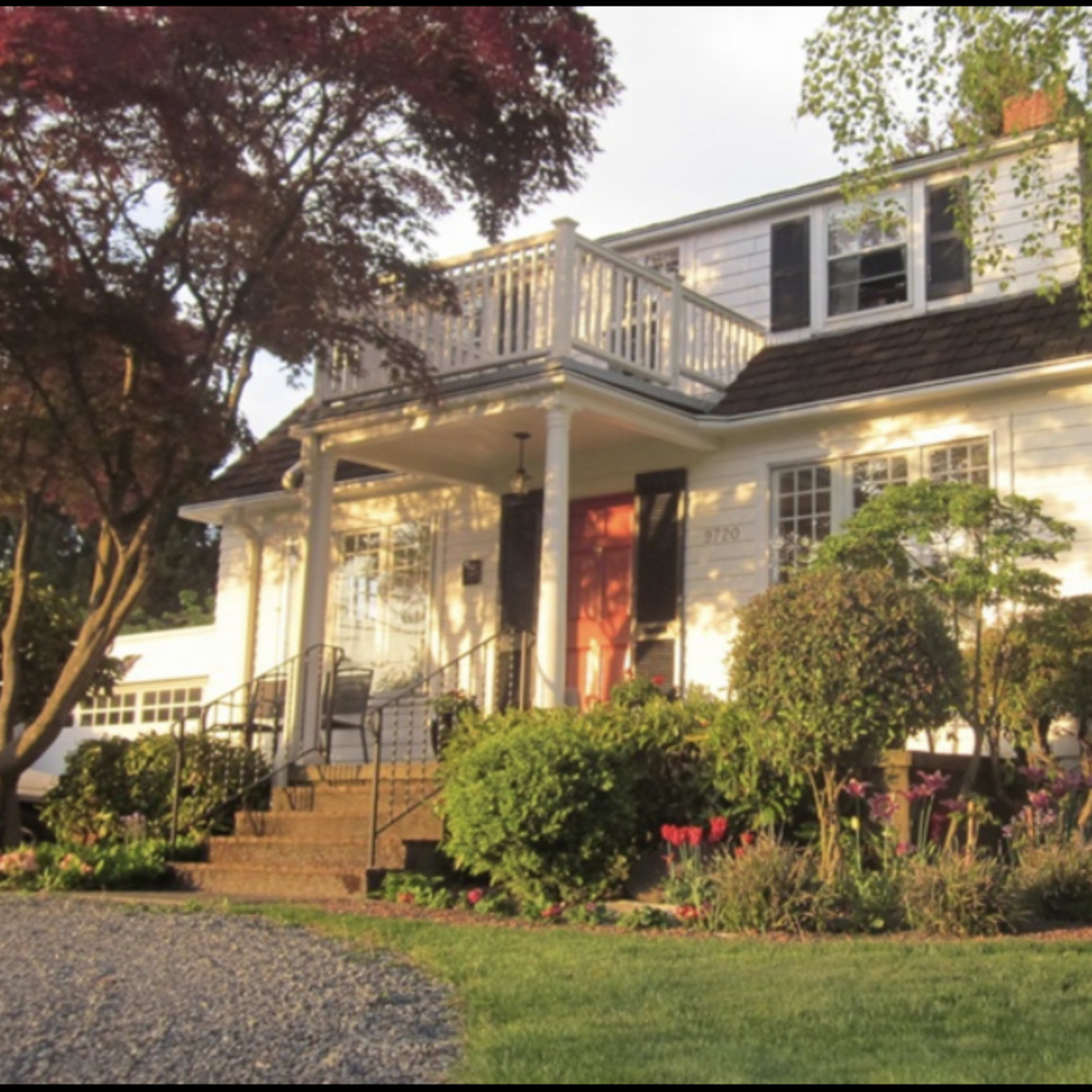 A two-story white house with a sloped roof, a front porch, and colorful garden landscaping.