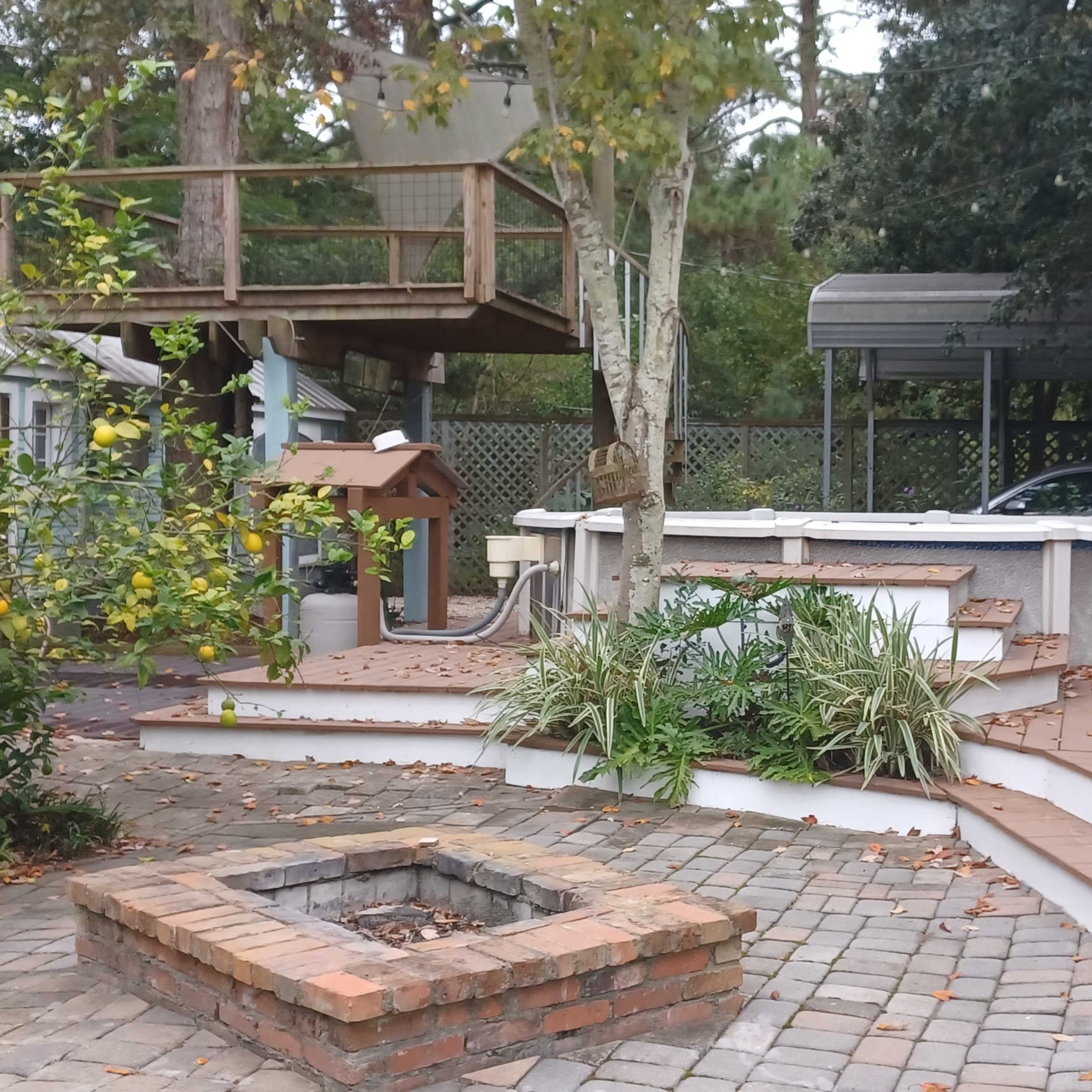 A landscaped backyard featuring a stone fire pit, a deck with a seating area, and a raised platform surrounded by trees and a carport.
