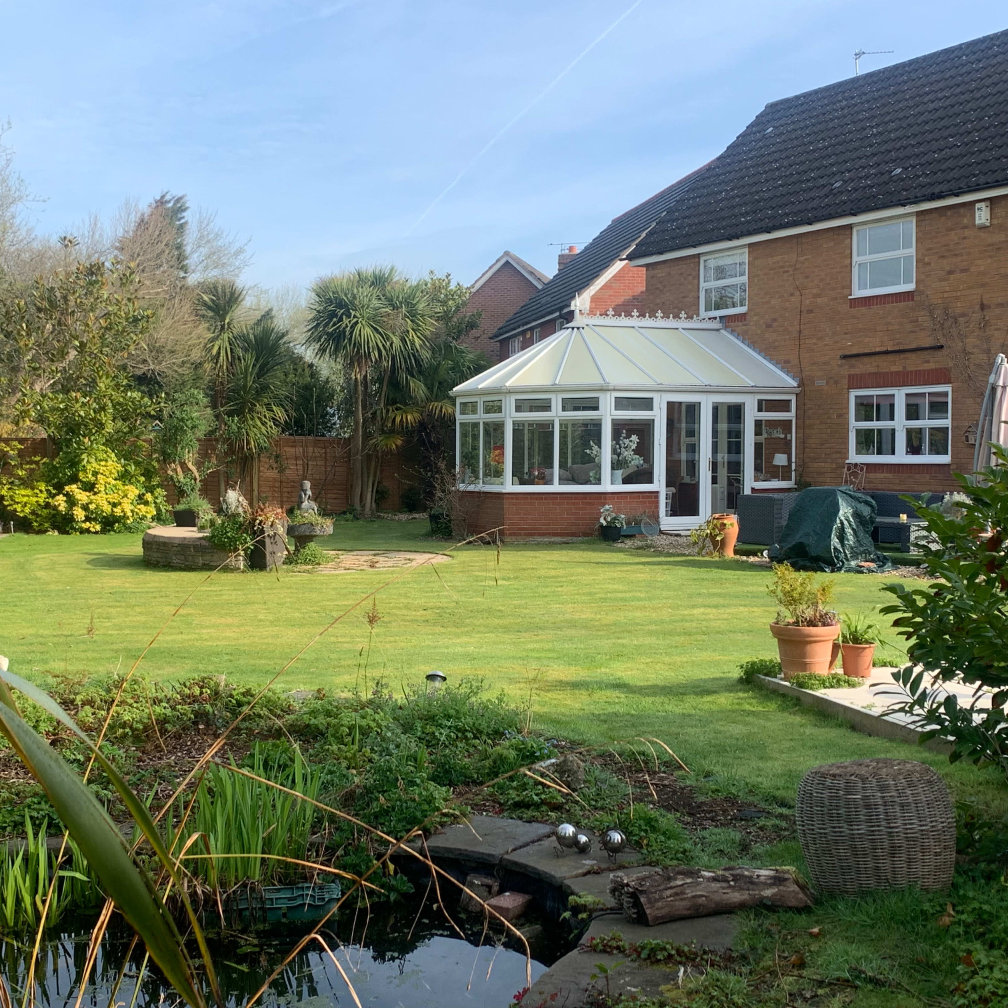 A well-maintained backyard featuring a grassy area, a conservatory, various plants, and a small pond.