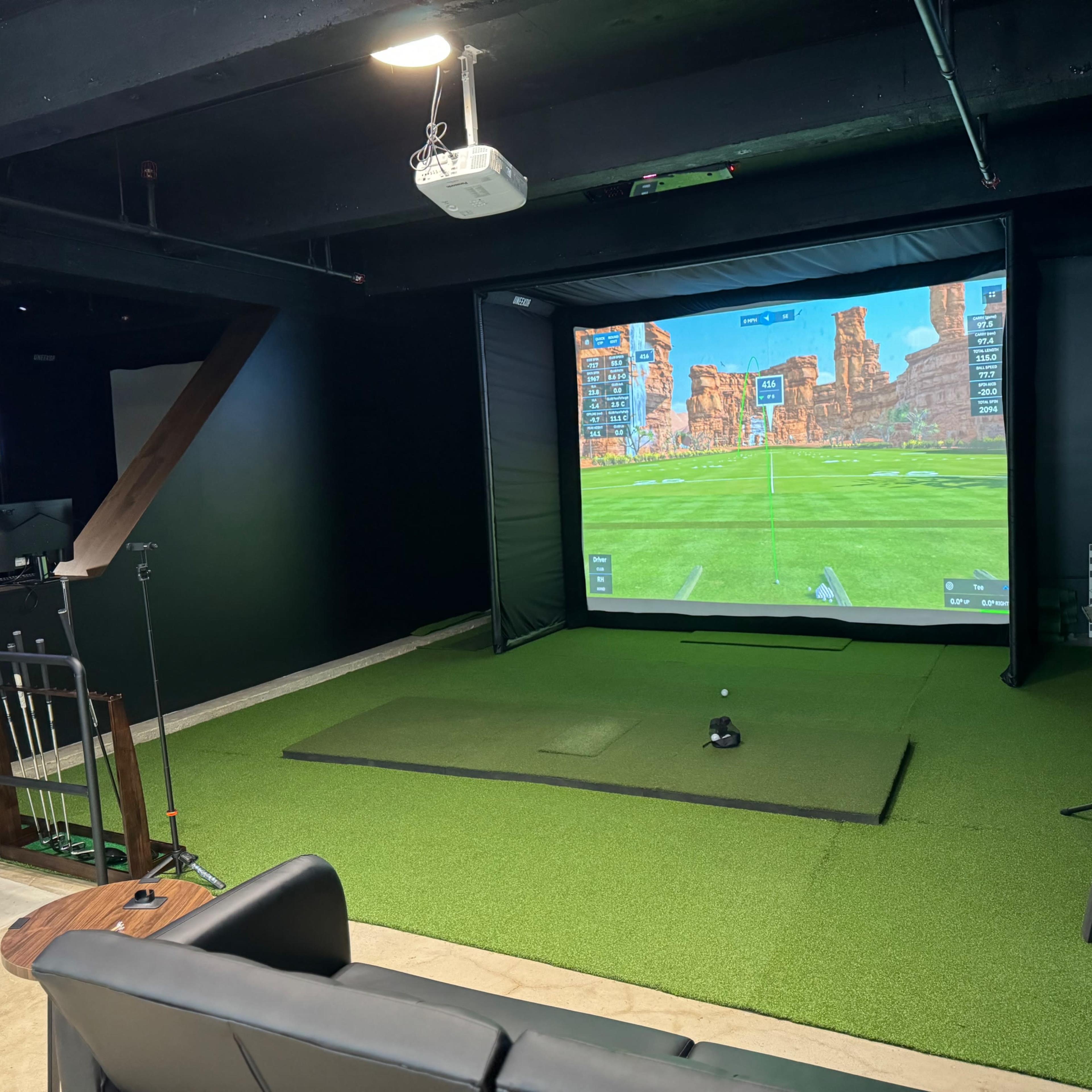 A golf simulator is set up in a room with a large screen displaying a golf course while a television shows a sports game in the background.