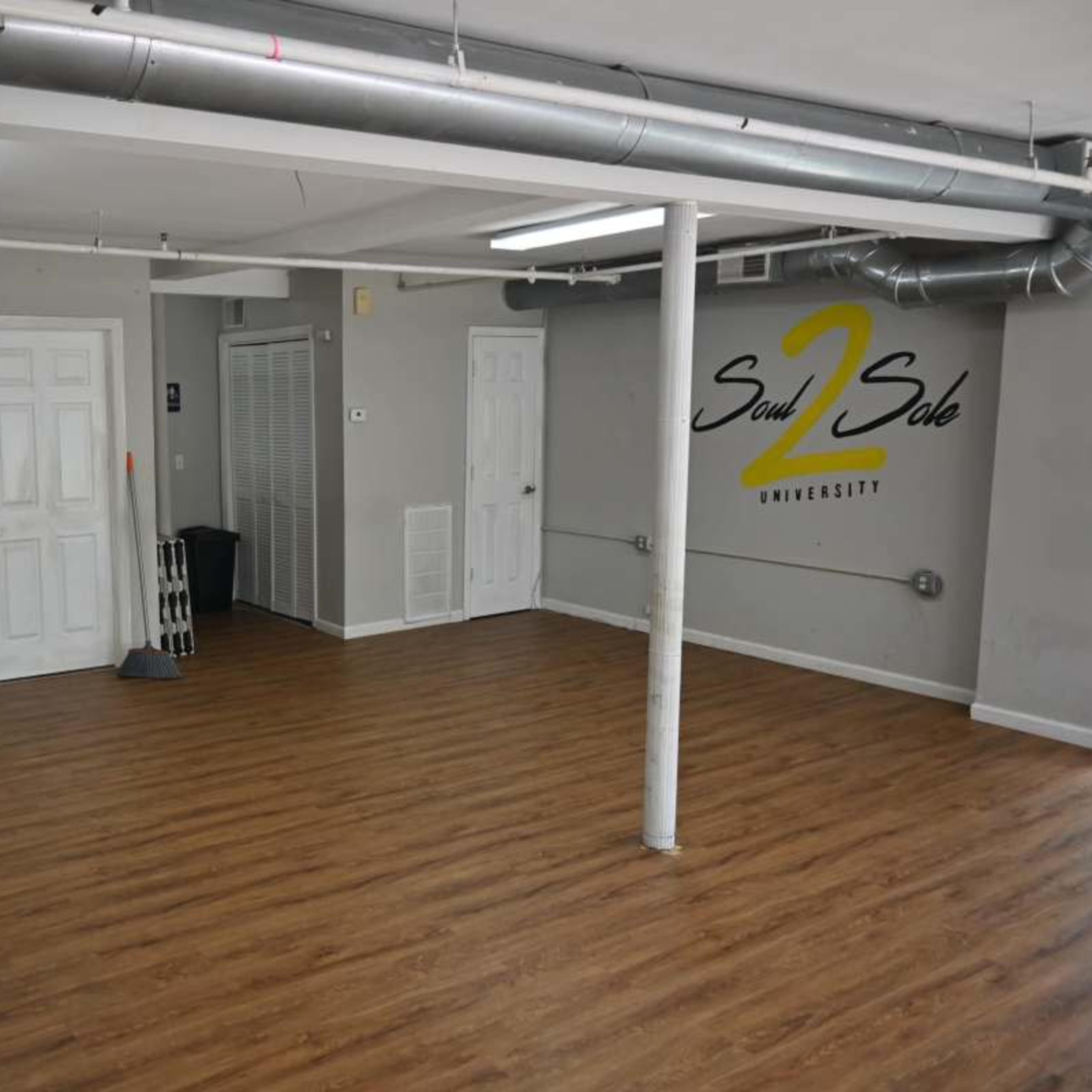 A spacious, well-lit room with wooden flooring, featuring a speaker system, a folding table, and the logo "Soul 2 Sole University" on the walls.