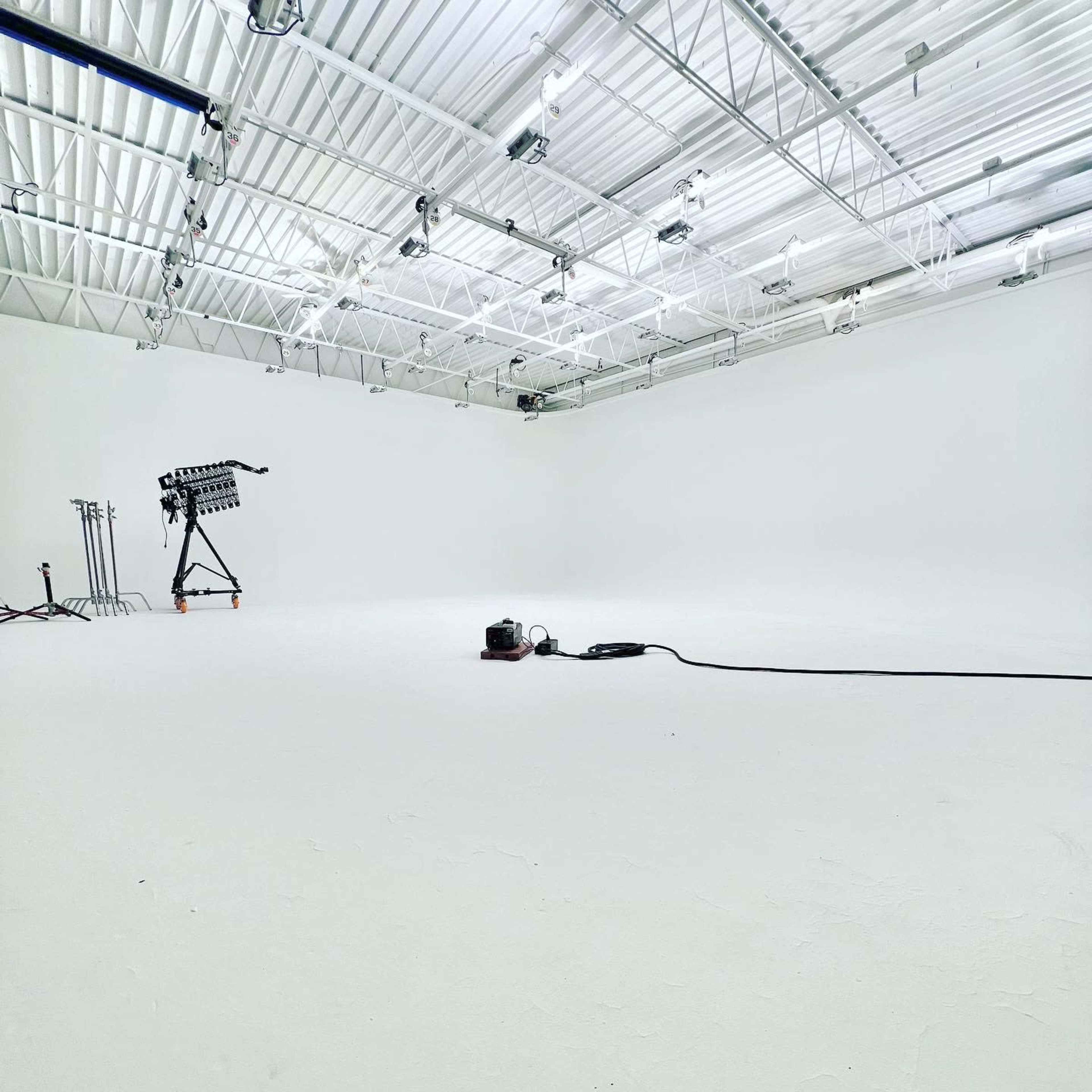 A spacious, empty studio with a white floor and walls, featuring lighting equipment on stands and a vacuum cleaner on the floor.
