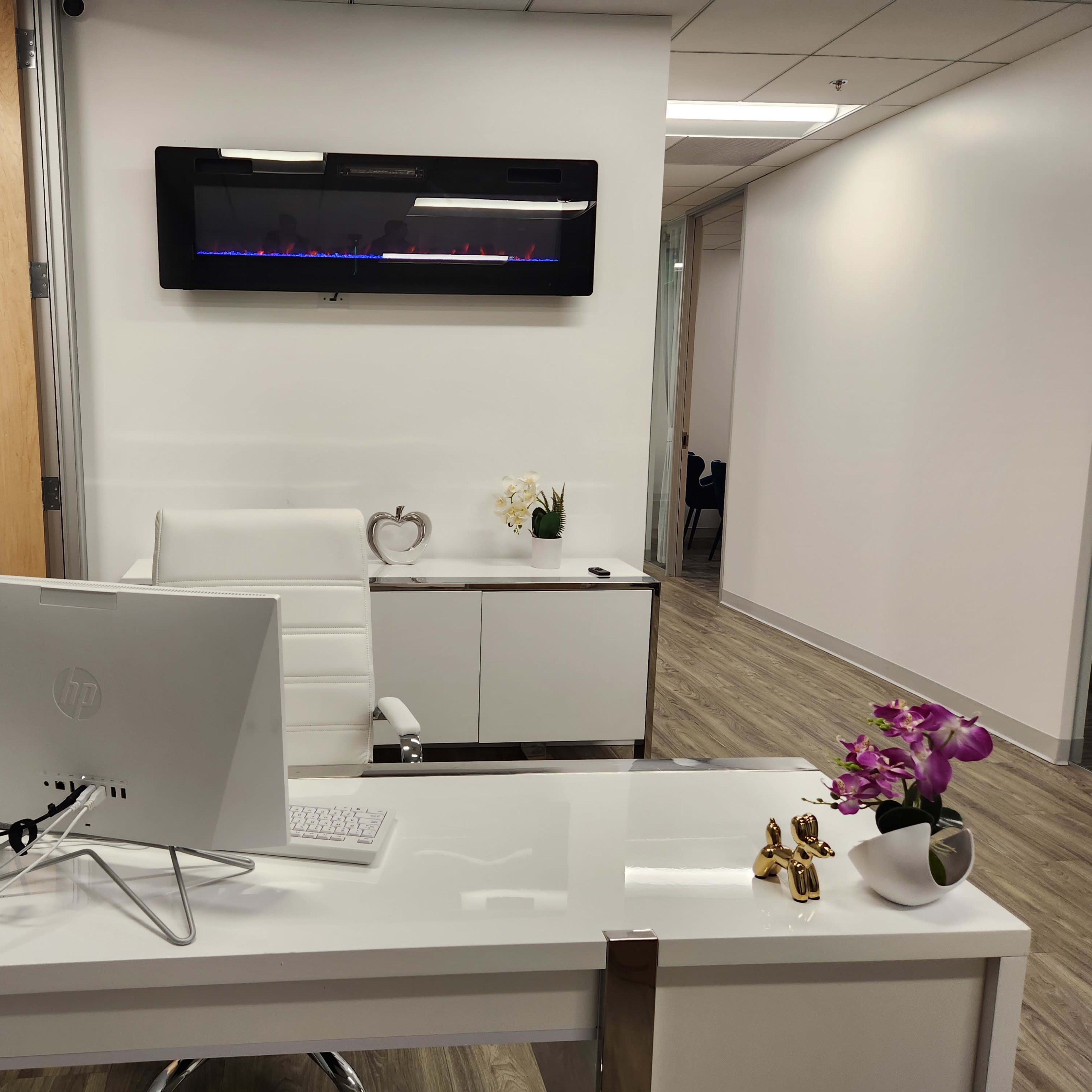 A modern office space featuring a white desk with a computer, a sleek chair, and a wall-mounted electric fireplace above a cabinet with decorative items.