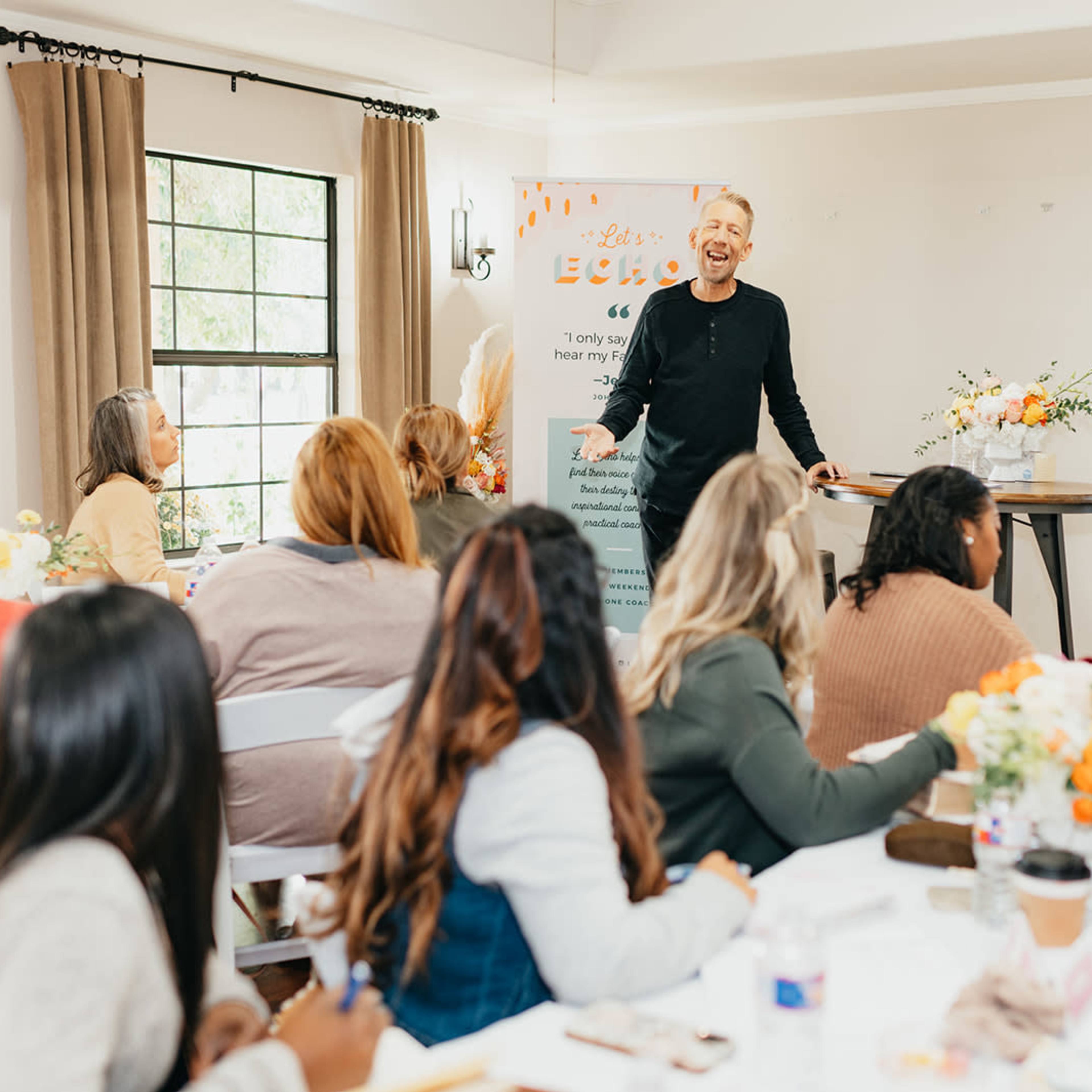 A speaker stands in front of an audience in a well-lit room, discussing ideas while participants take notes at tables adorned with floral centerpieces.