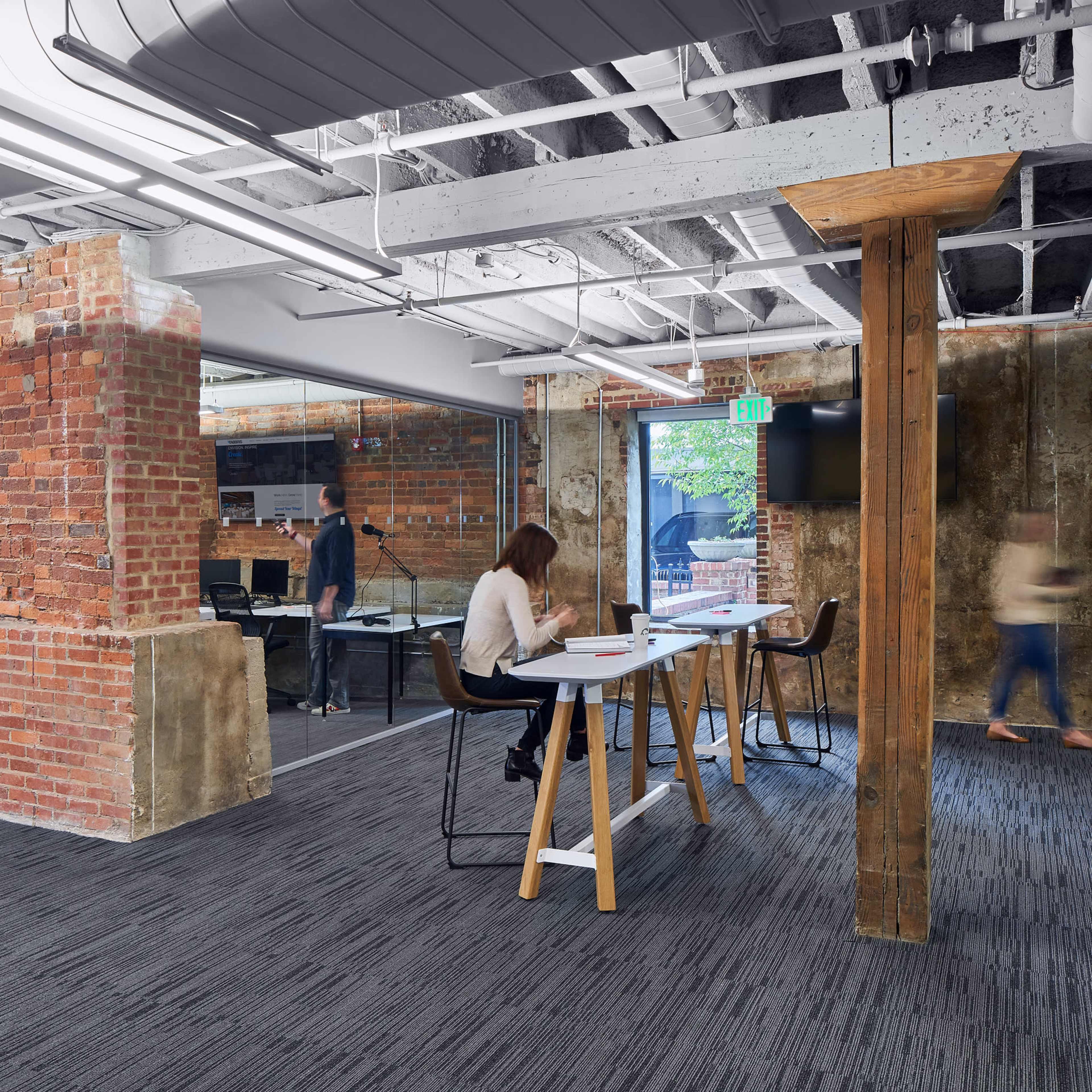 An open office space with exposed brick walls, wooden columns, and modern furniture, where several people are working and walking around.