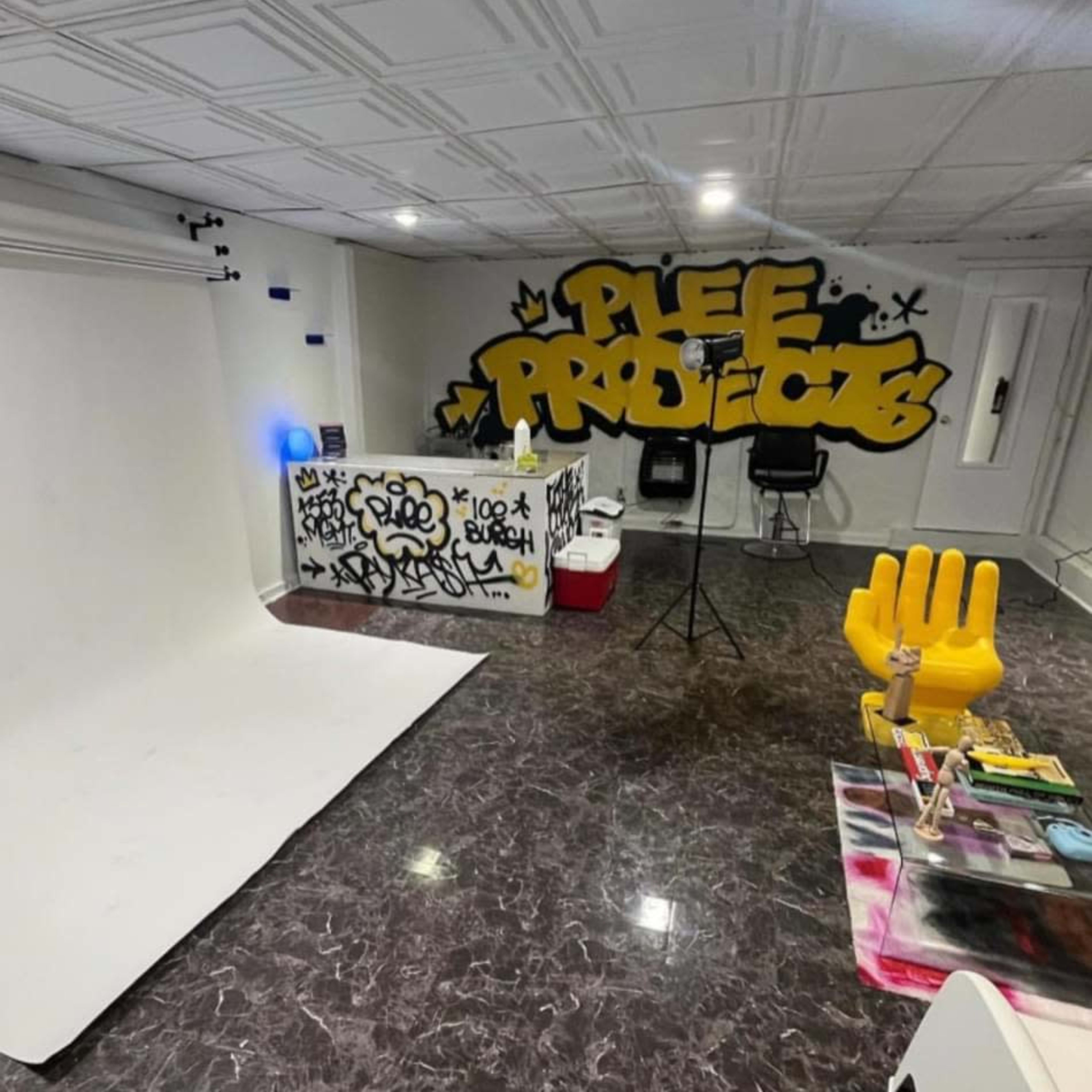 A studio space with a graffiti wall featuring the words "Plee Projects," a stark white backdrop, and various artistic supplies arranged on a table.