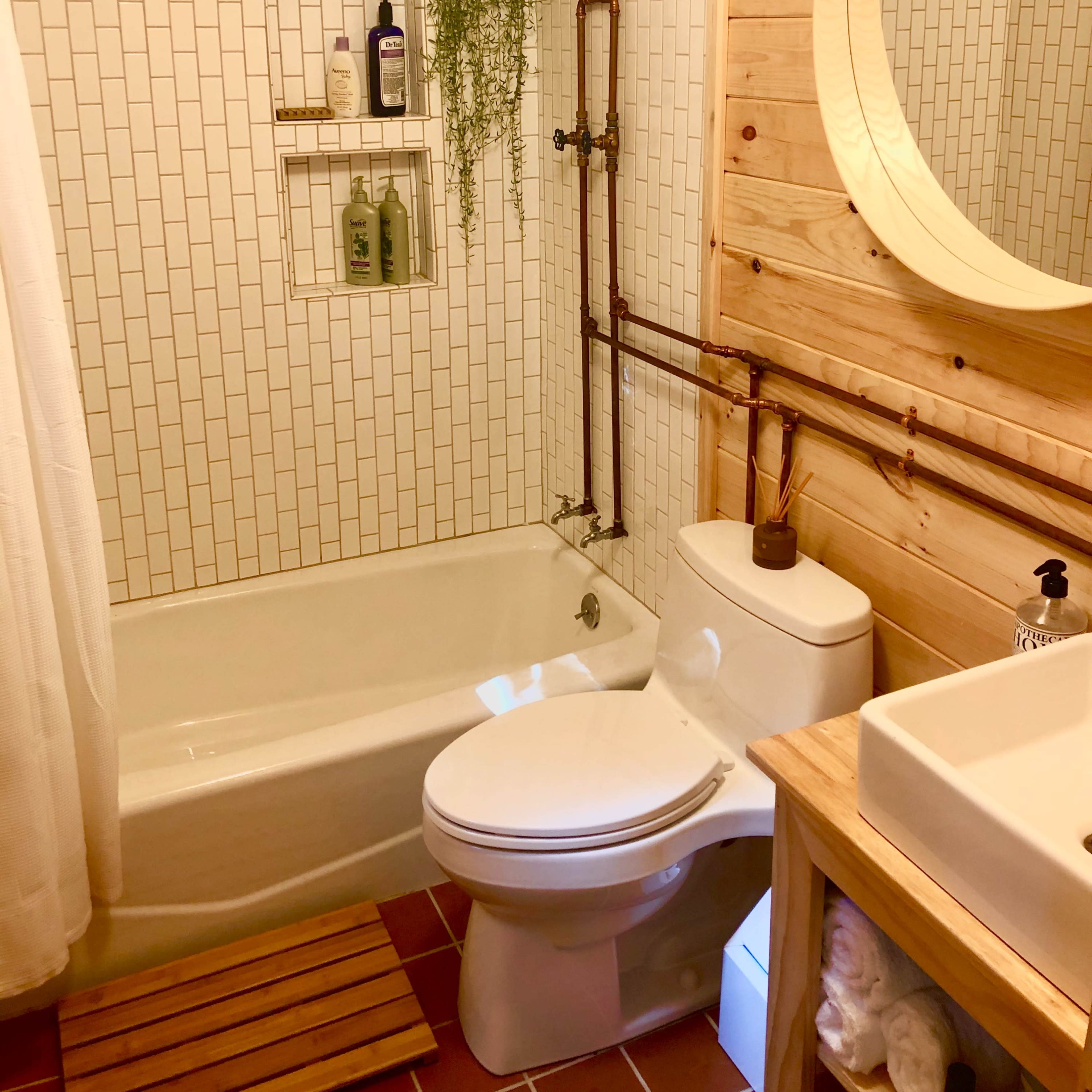 A small bathroom featuring a bathtub, a wall-mounted toilet, and a sink with wooden cabinetry, all set against a backdrop of white tiled walls and natural wood accents.