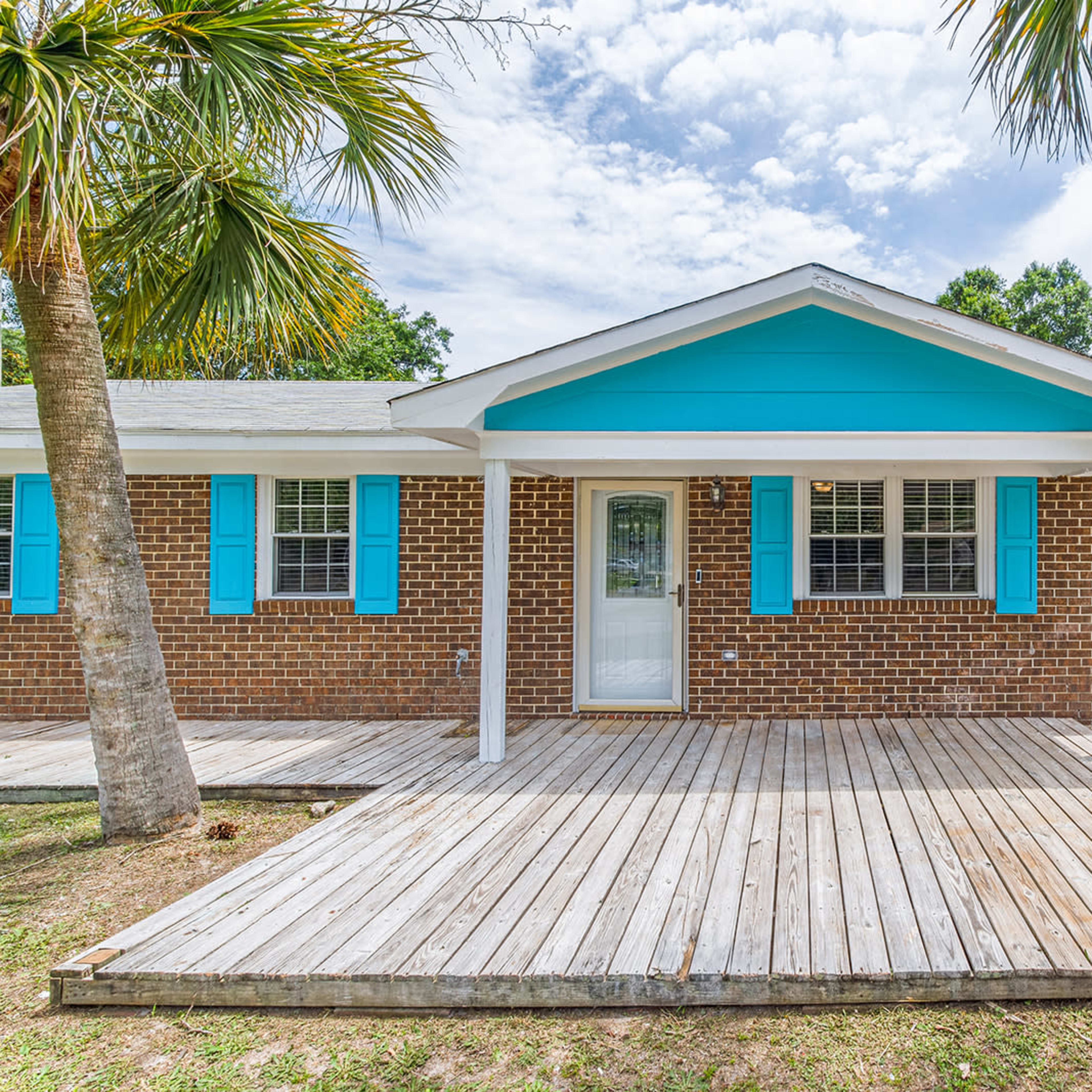 A brick house with turquoise shutters, a covered porch, and a wooden deck surrounded by palm trees.