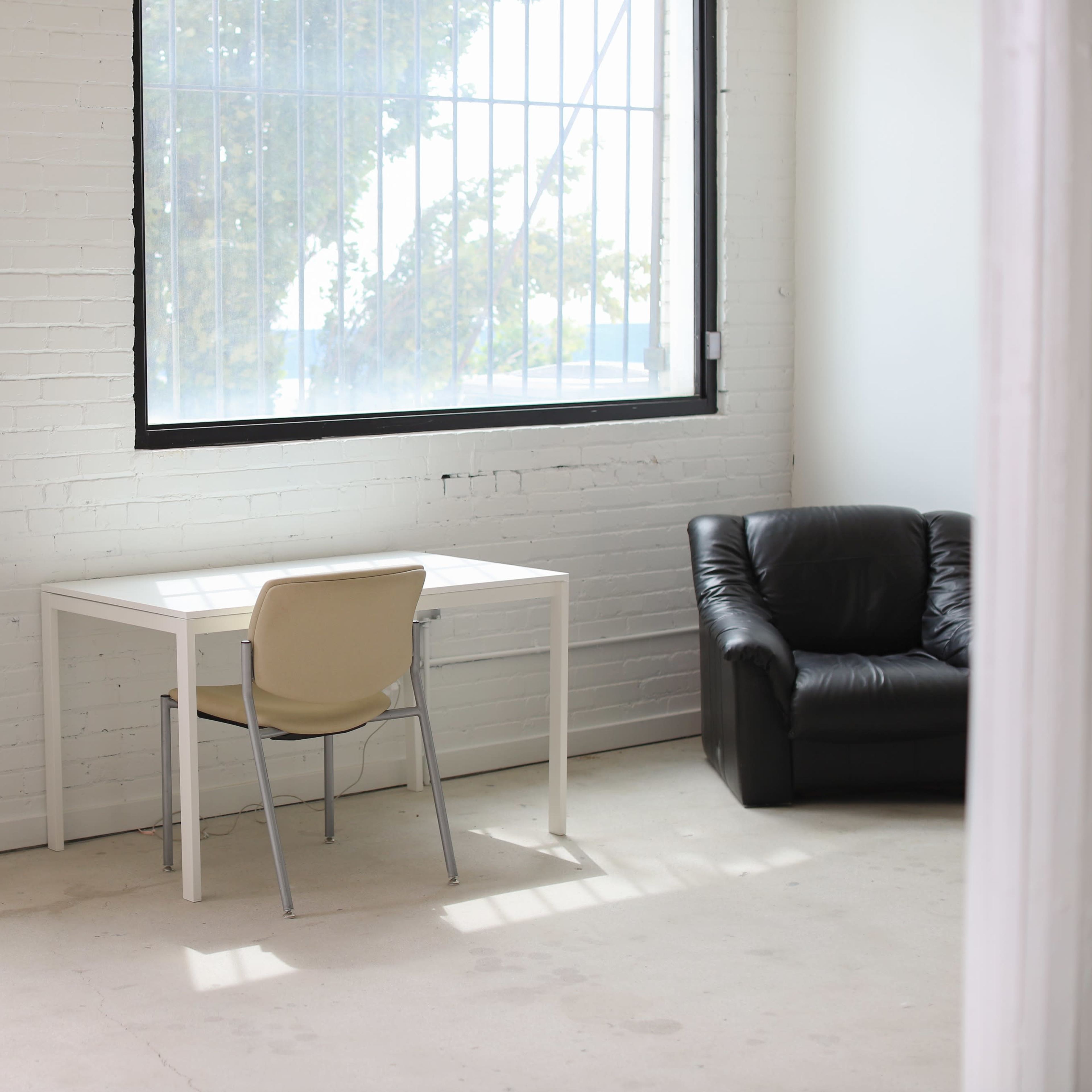 A modern room features a large mirror, a white desk with a chair, and a black leather armchair beside a window.