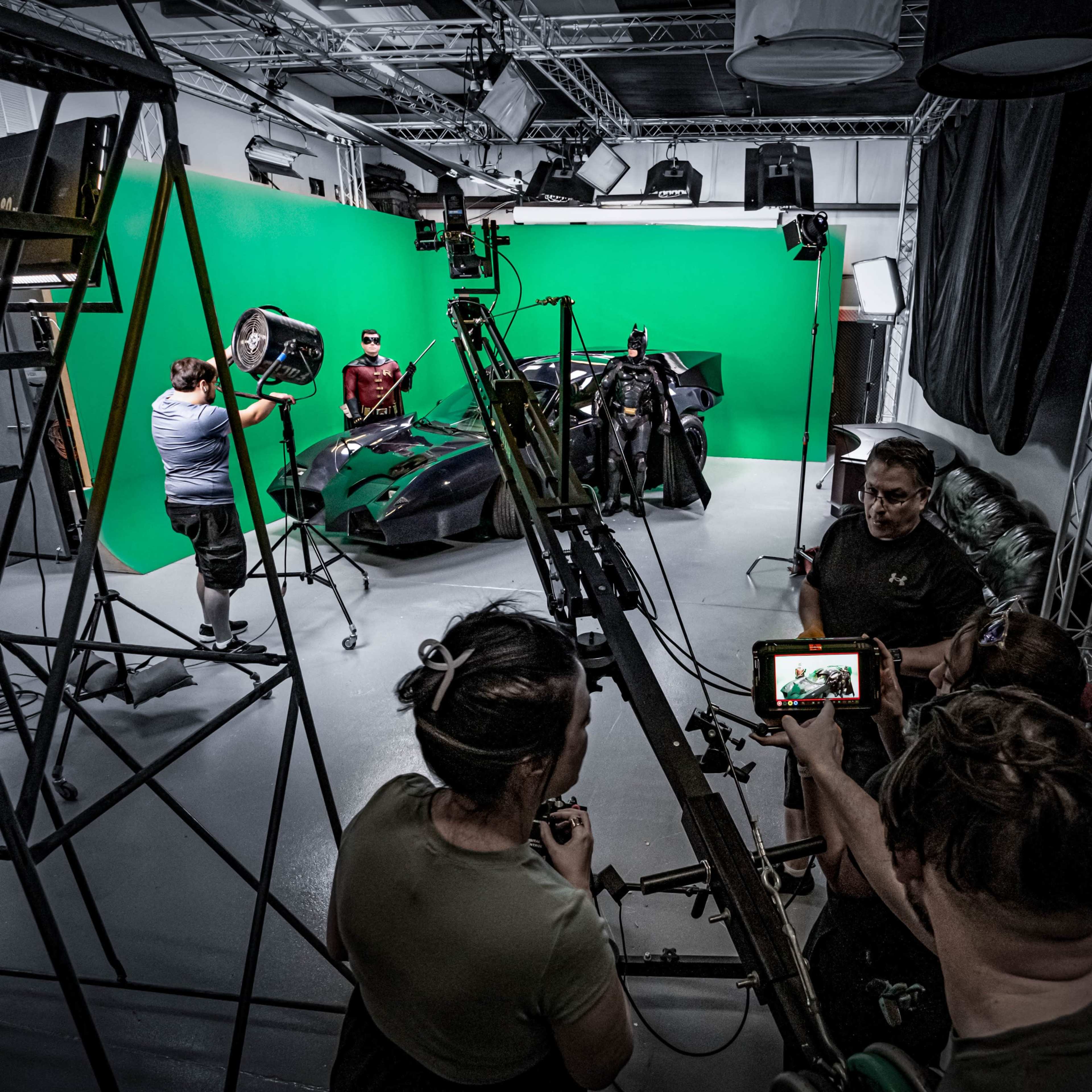 A film crew sets up a scene in a studio with a green screen, featuring a Batmobile and actors in costume.
