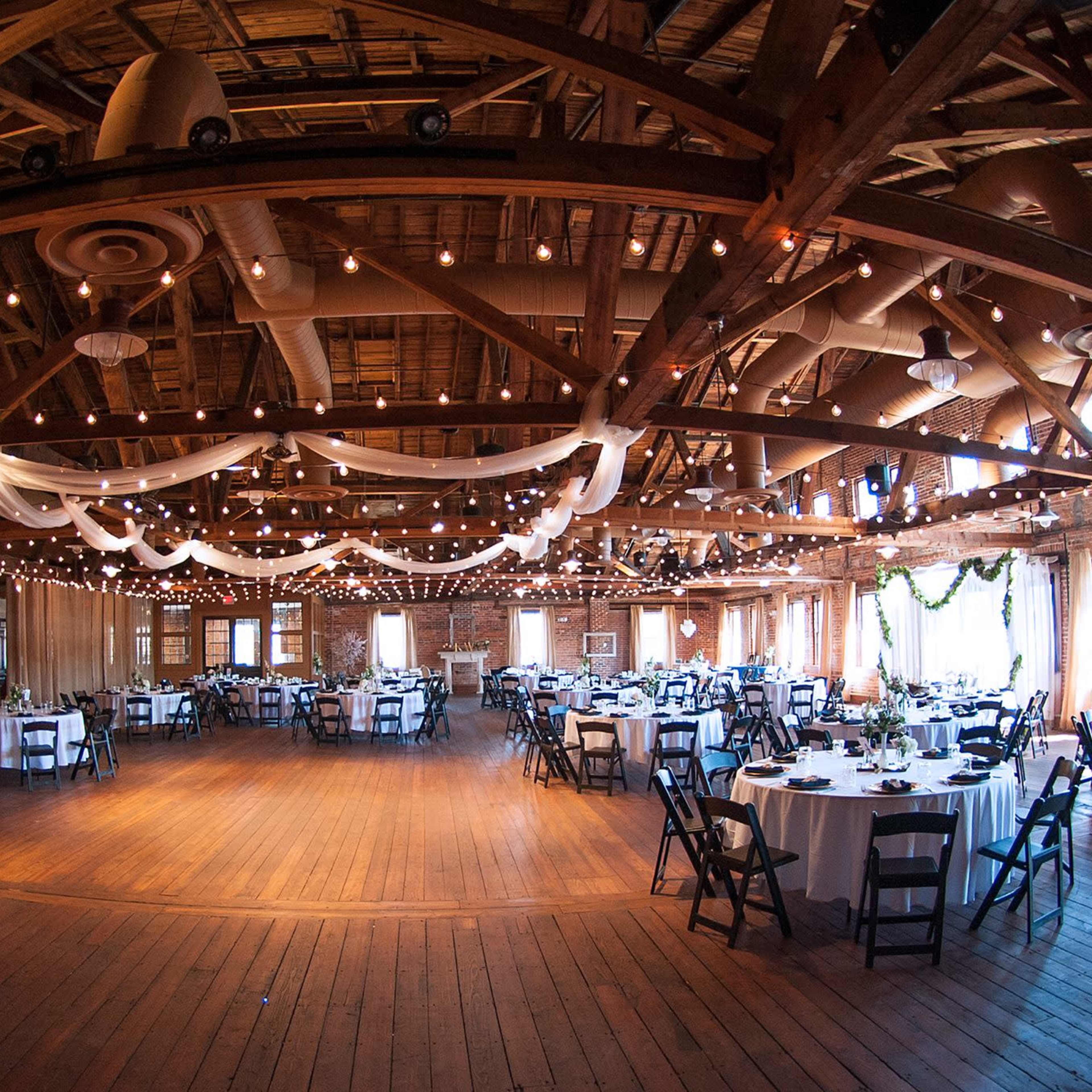 A large event space with multiple round tables covered in white tablecloths, set up under string lights and draped fabric.