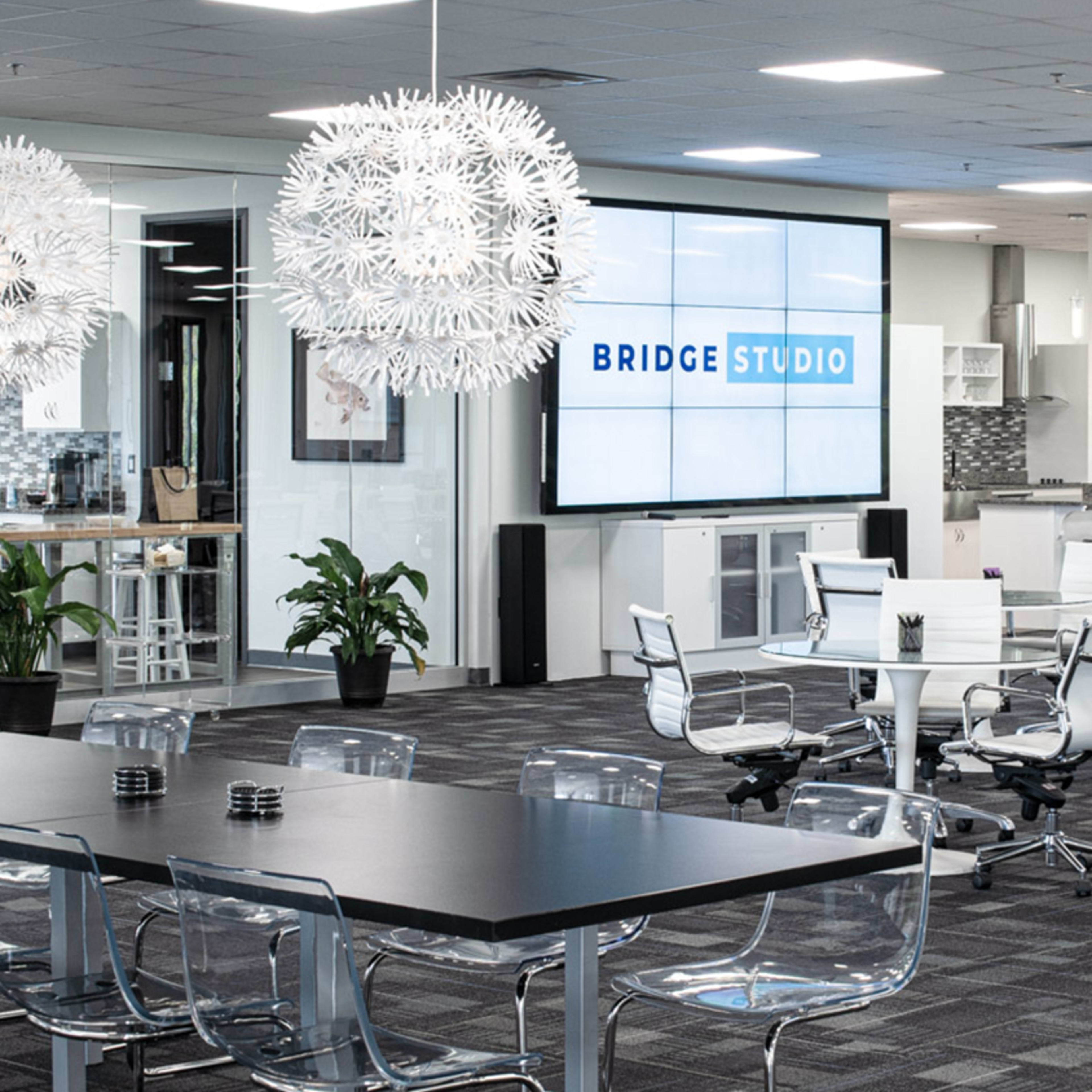A modern office space with a large screen displaying "BRIDGE STUDIO," surrounded by transparent chairs and a kitchen area in the background.