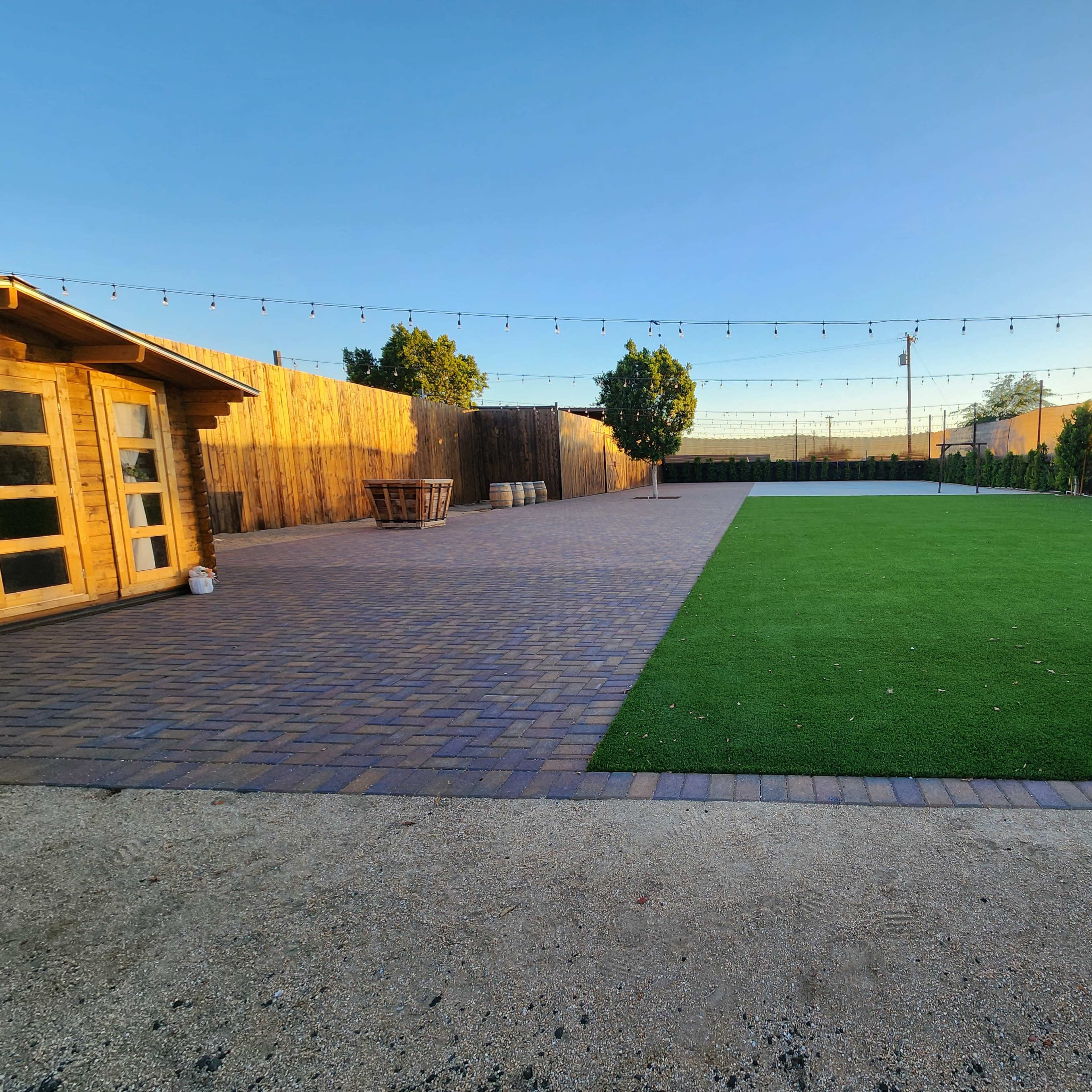 A spacious outdoor area featuring a section of artificial grass, a paved pathway, a wooden shed, and string lights above, with trees lining the perimeter.
