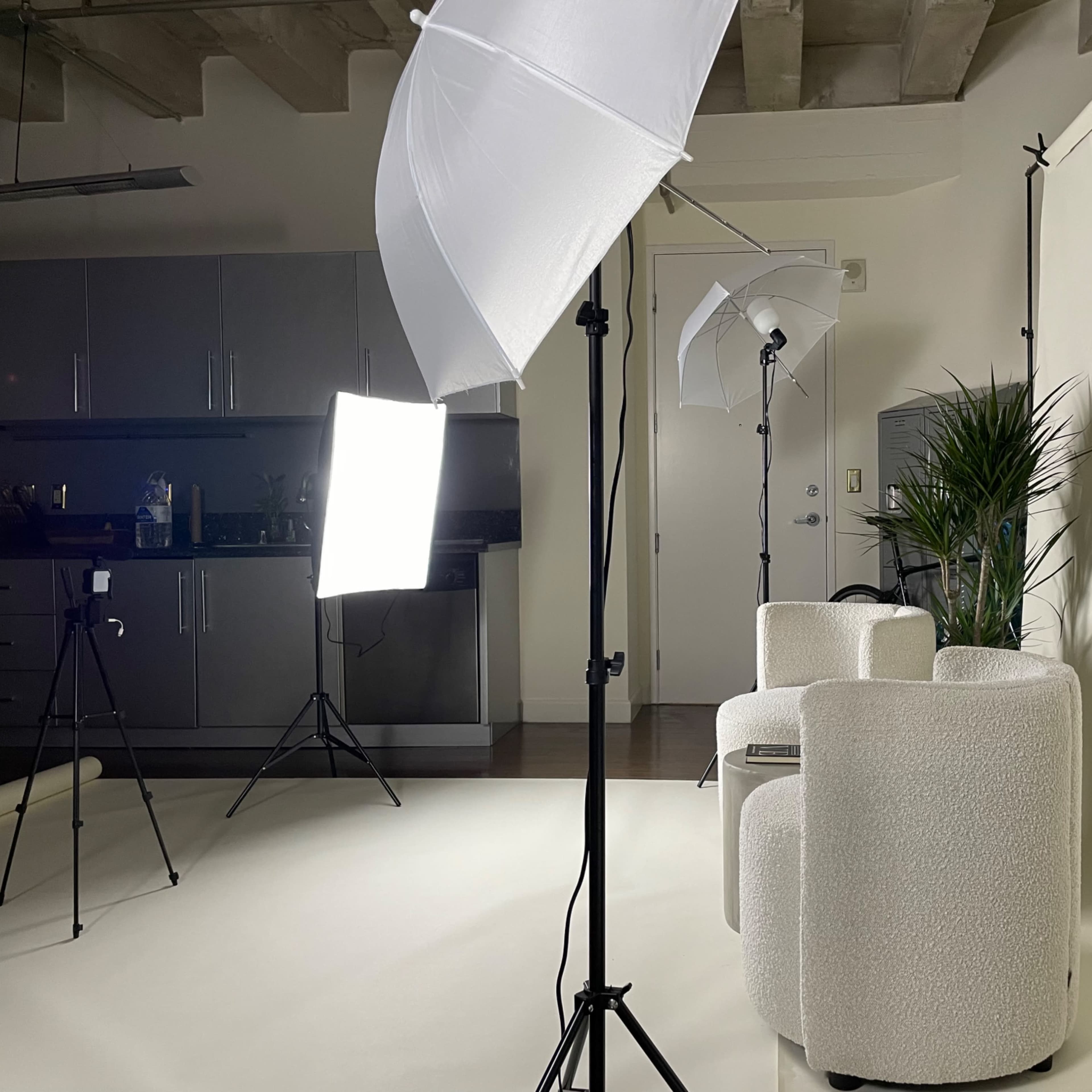 A photography studio setup with two light sources, including umbrella lights, and a minimalist seating arrangement against a plain backdrop.