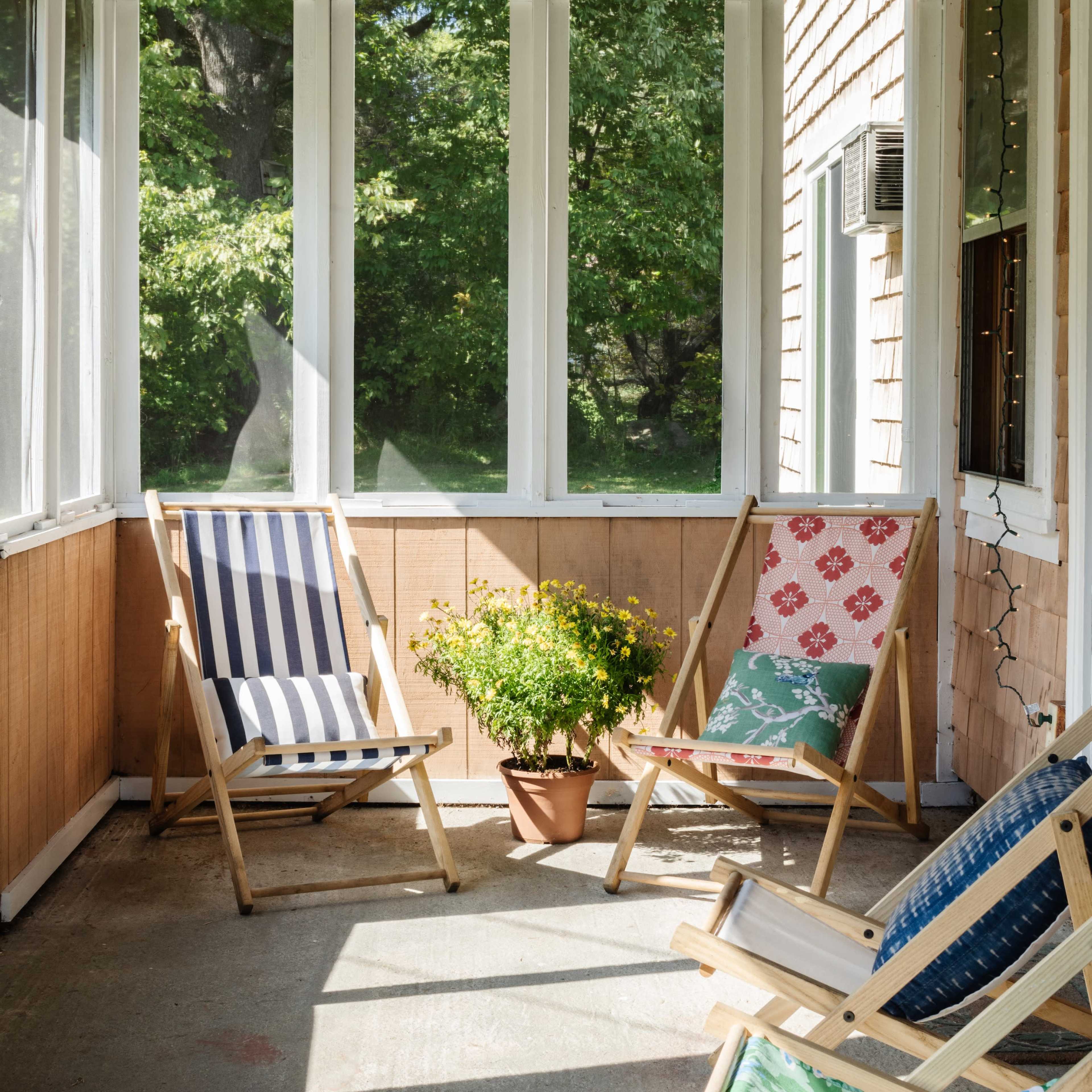 A sunlit screened porch features several wooden folding chairs and a potted plant on a concrete floor.