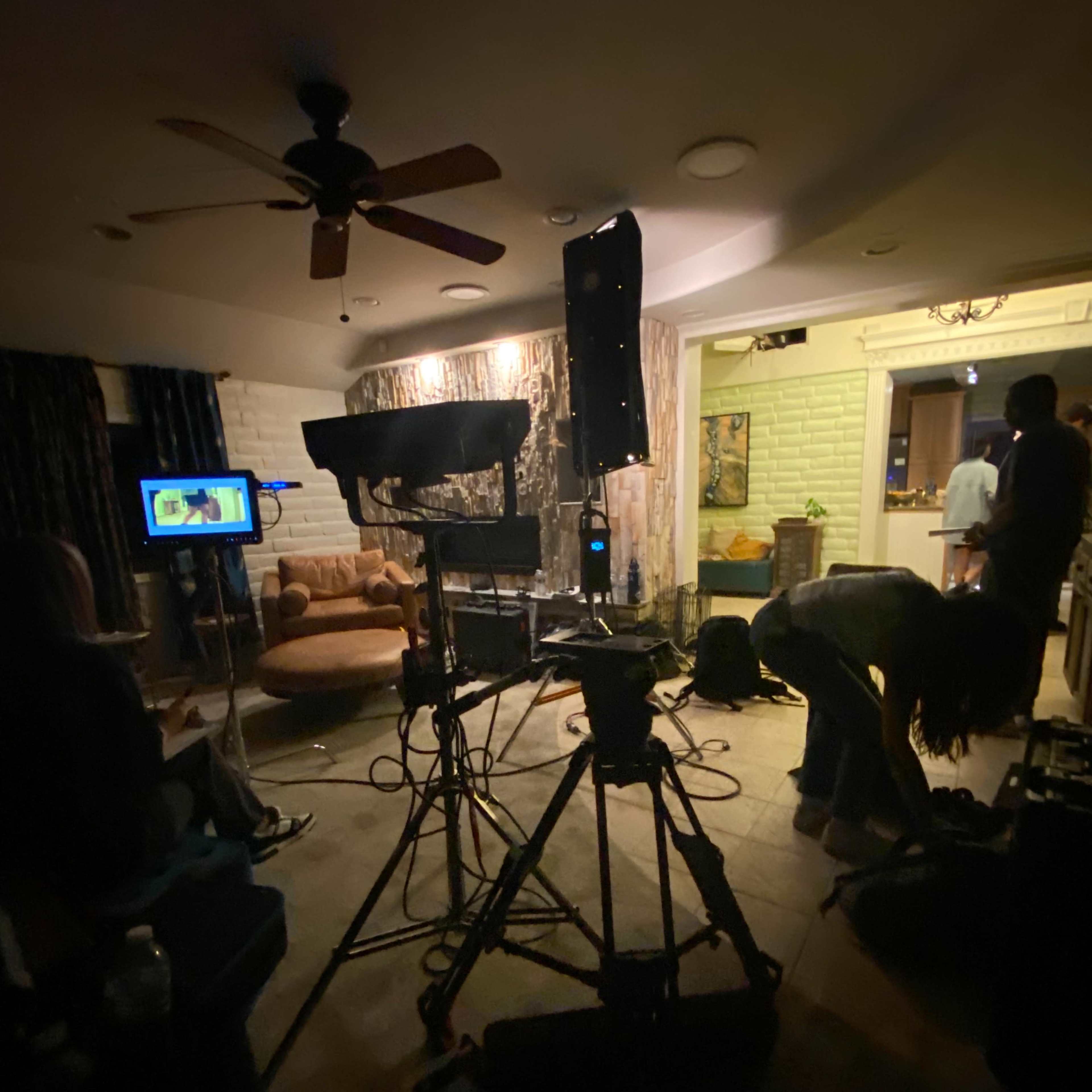 A film crew sets up camera equipment in a dimly lit room with a couch and a television display.