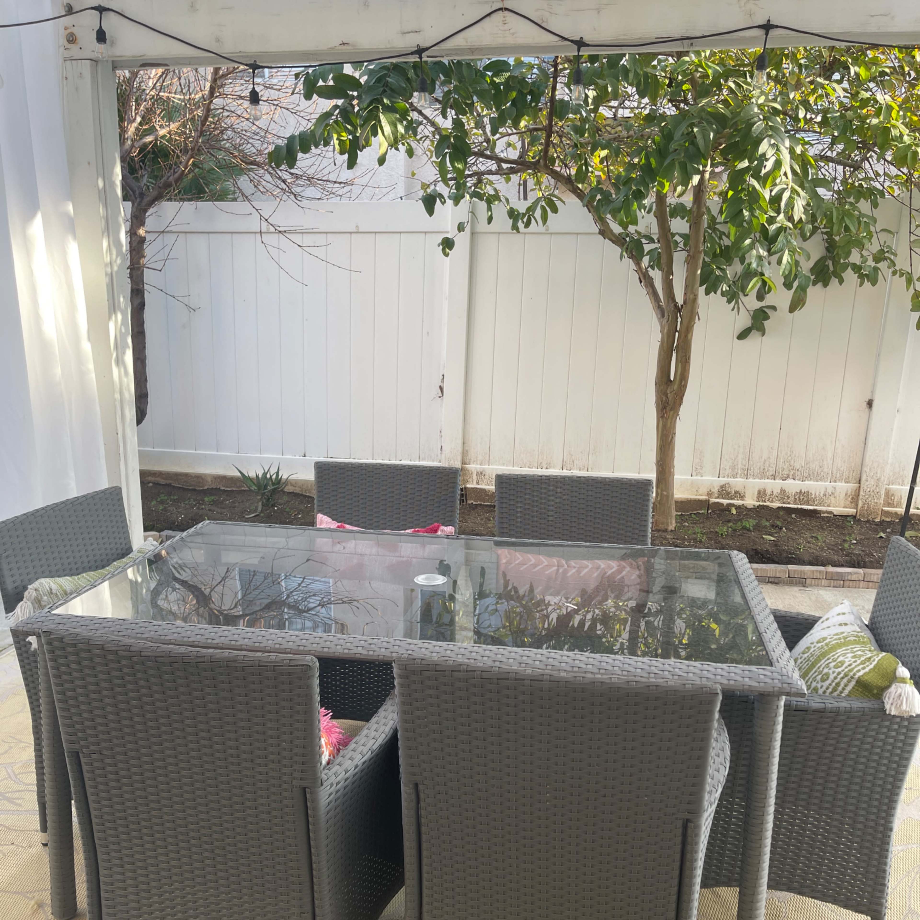 A rectangular glass table is surrounded by six wicker chairs on a patio under a covered structure, with a tree and fence in the background.