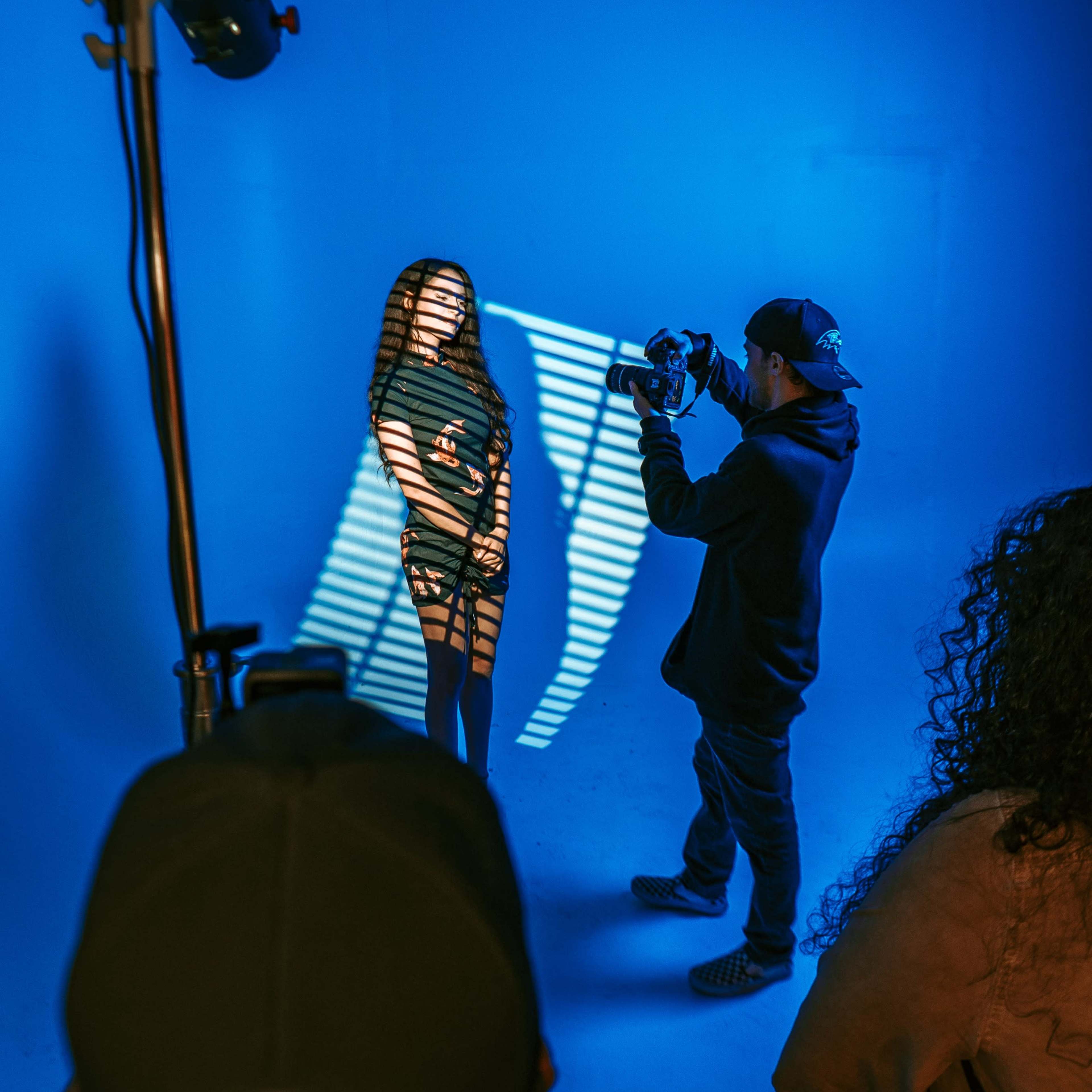 A photographer captures an image of a model standing under blue lighting, with shadows casting stripes on her body from a nearby light source.