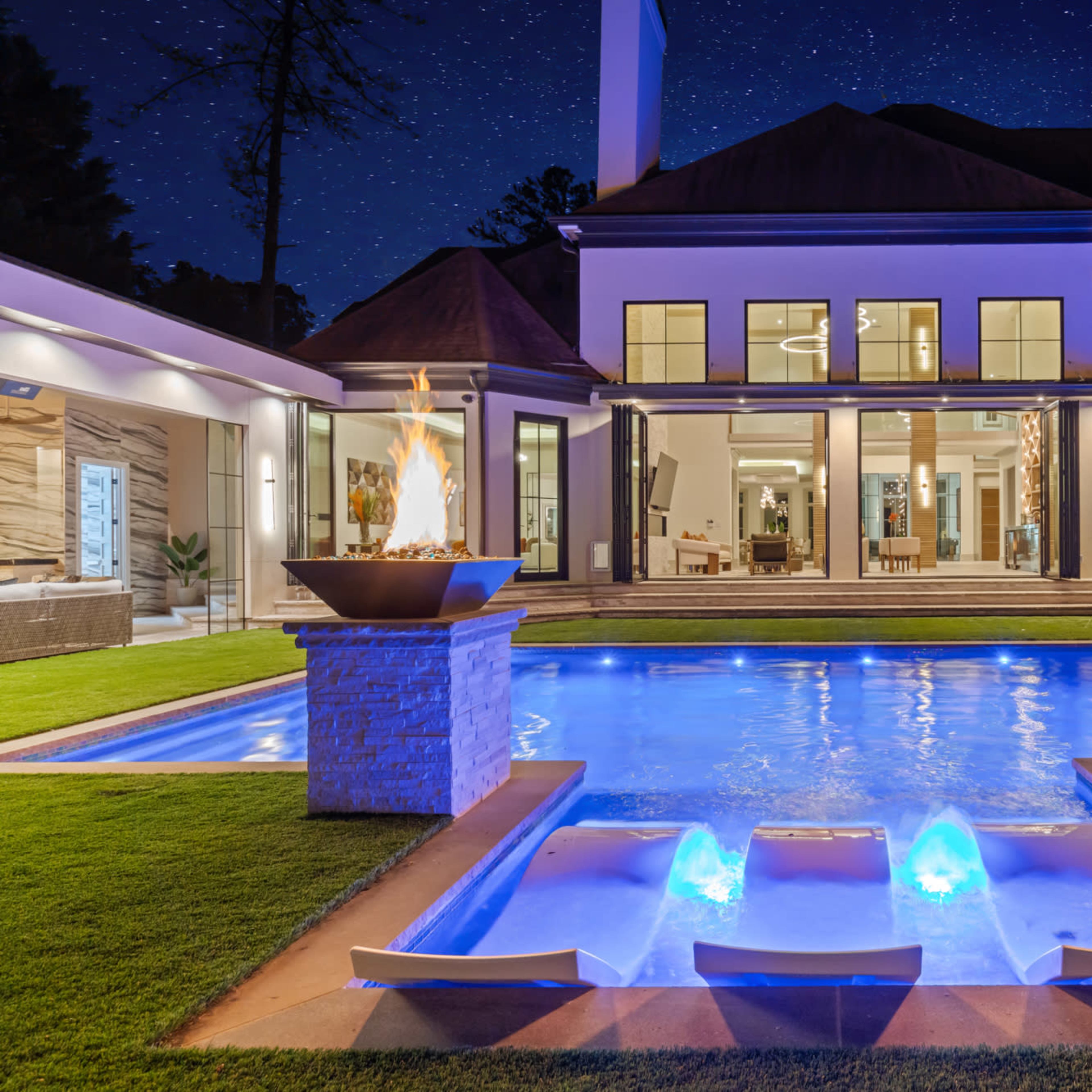 A brightly lit backyard features a swimming pool with water fountains, flanked by two fire bowls, and a modern house replete with large windows in the background.