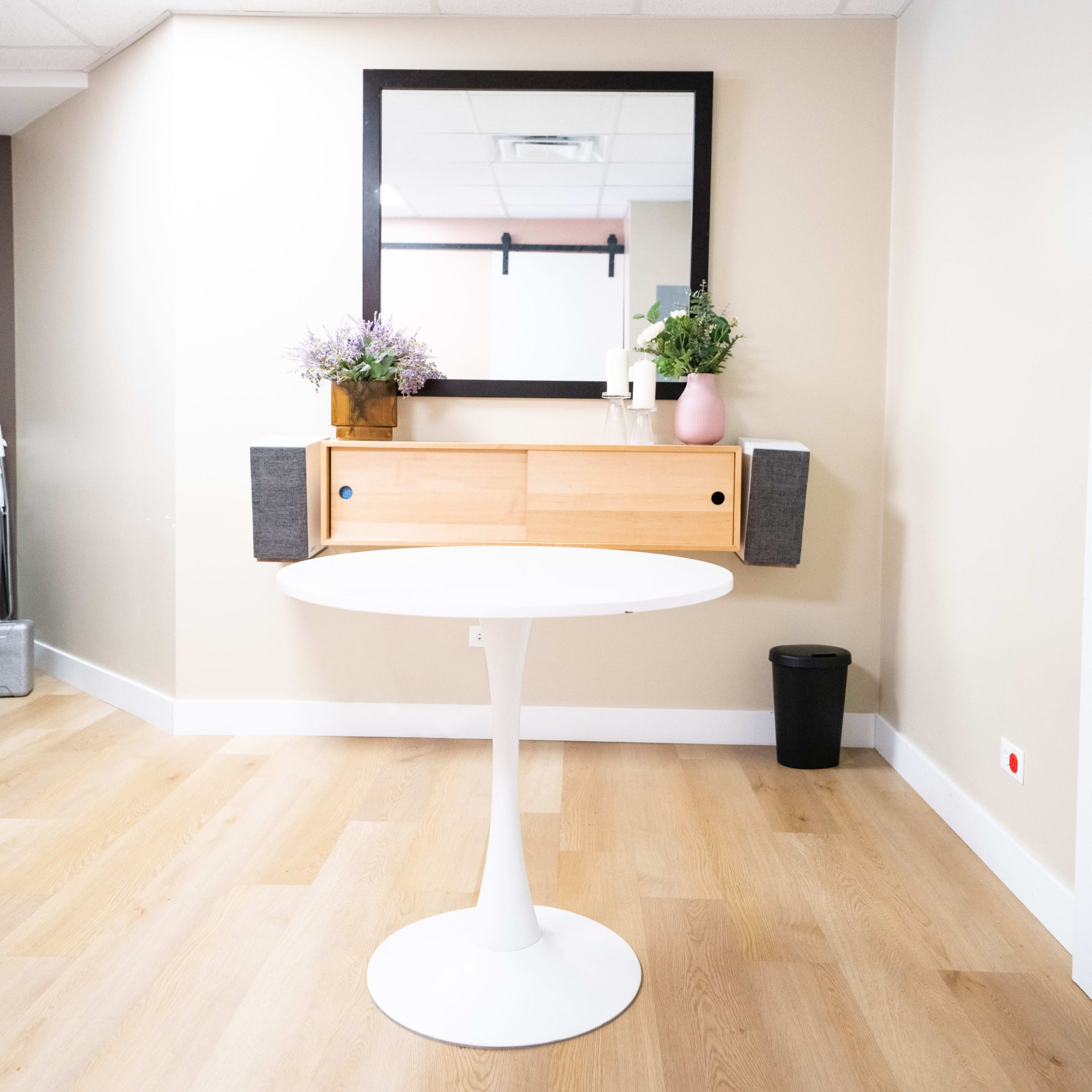 A round white dining table is centered in a room with wooden floors, featuring a wall-mounted mirror above a wooden console flanked by potted plants.