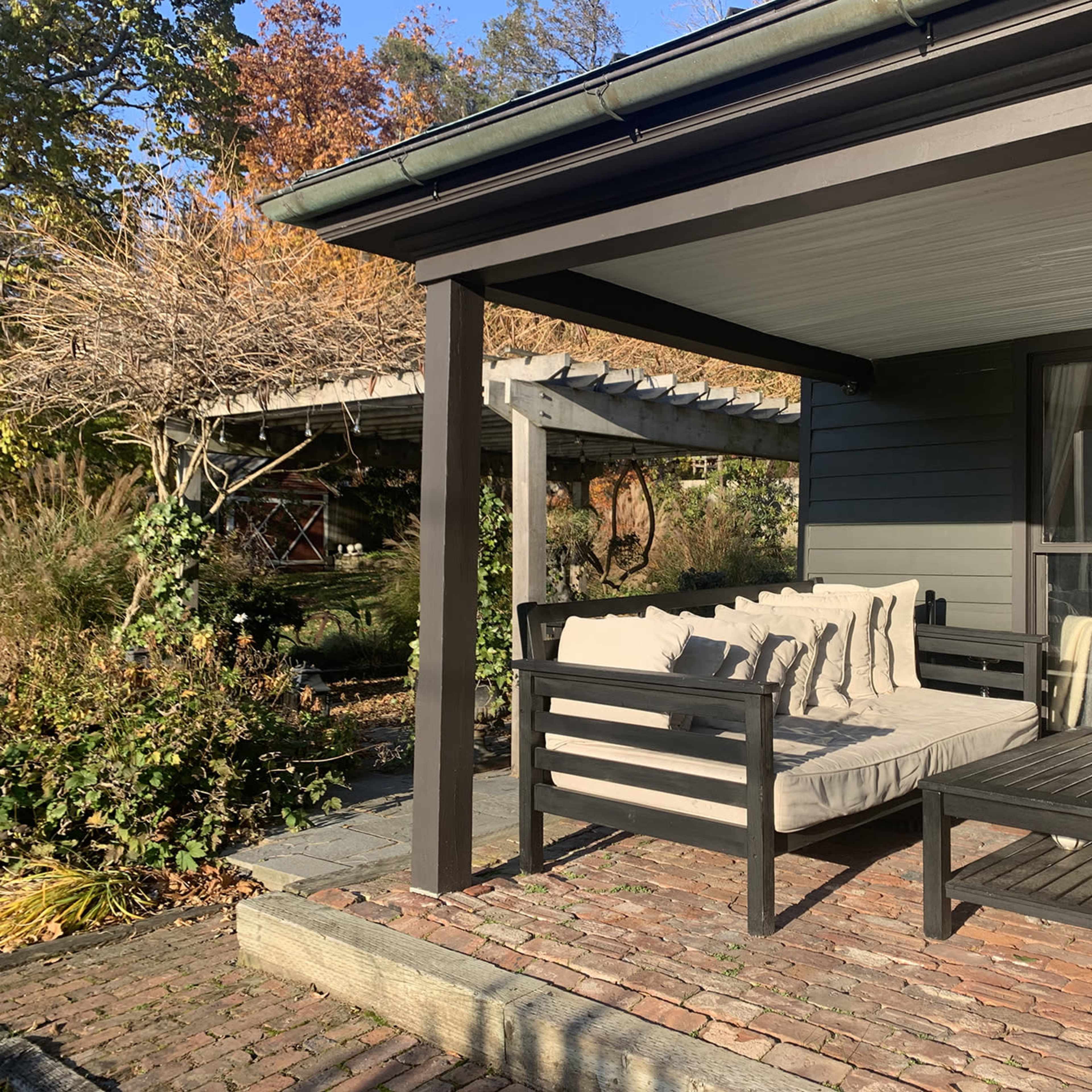 A patio area with a sofa and a coffee table, surrounded by greenery and a wooden structure in the background.