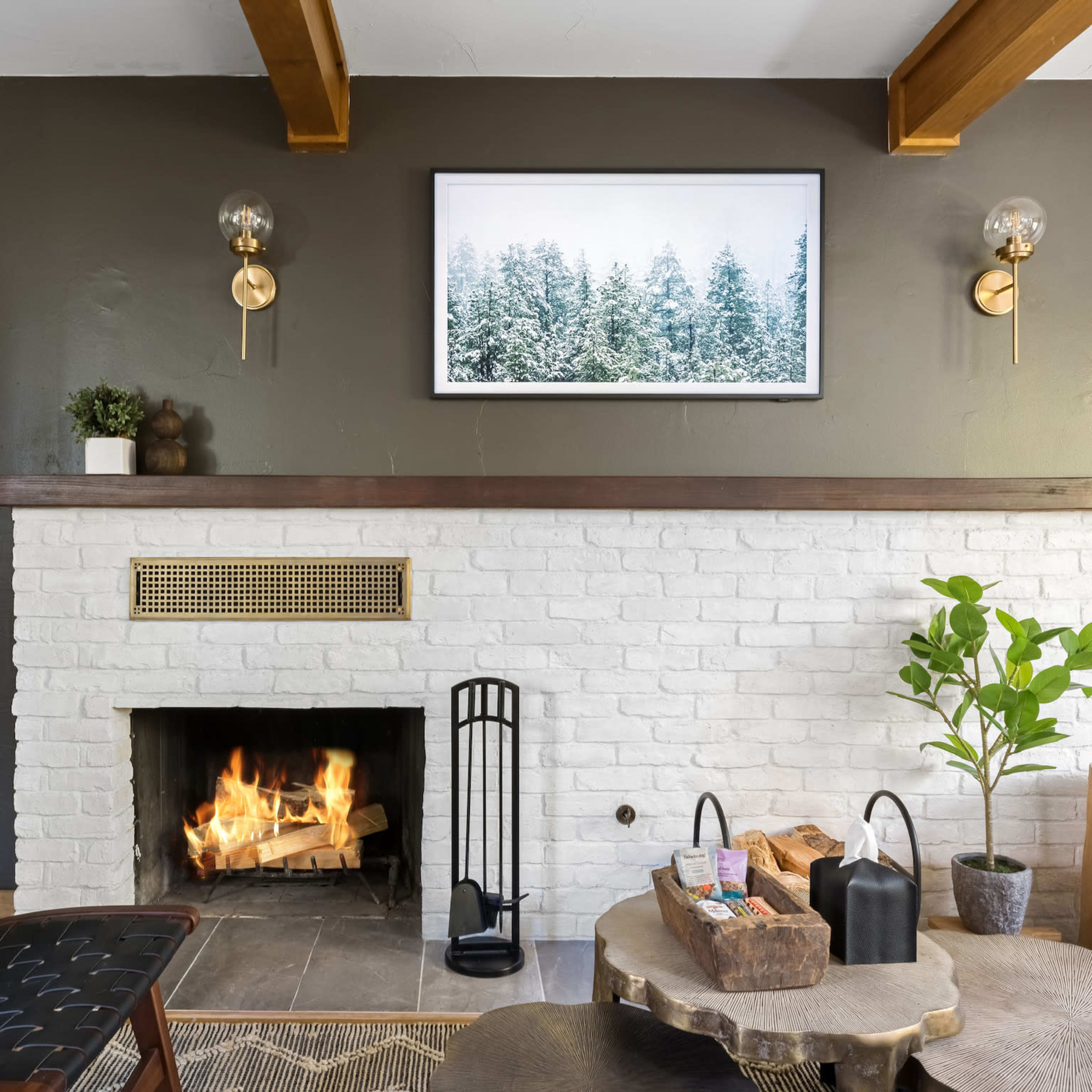 A cozy living room features a white brick fireplace with a fire burning, surrounded by modern decor elements including a potted plant and wall-mounted light fixtures.