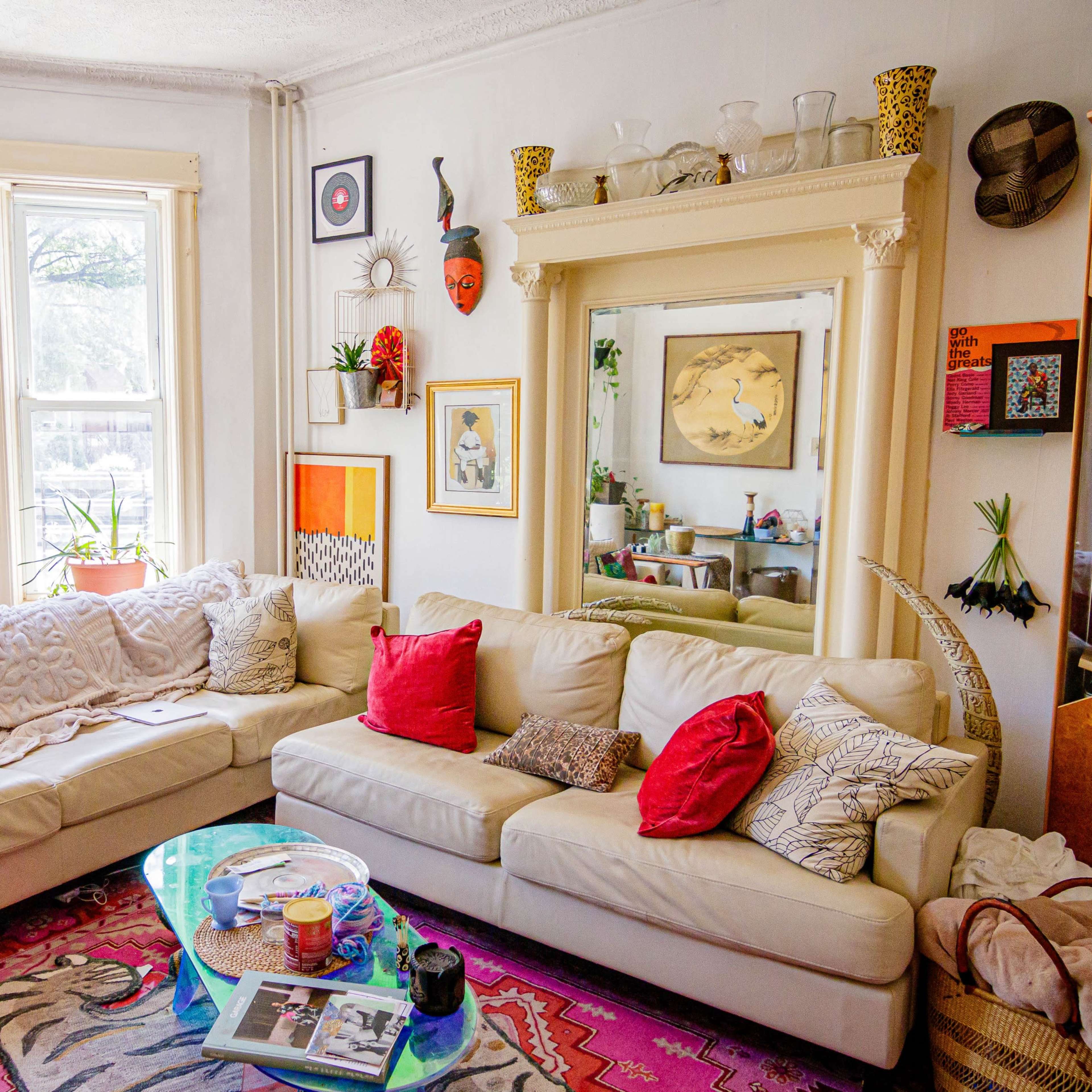 The living room features a sectional sofa, colorful cushions, a glass coffee table, and decorative plants positioned around large windows.