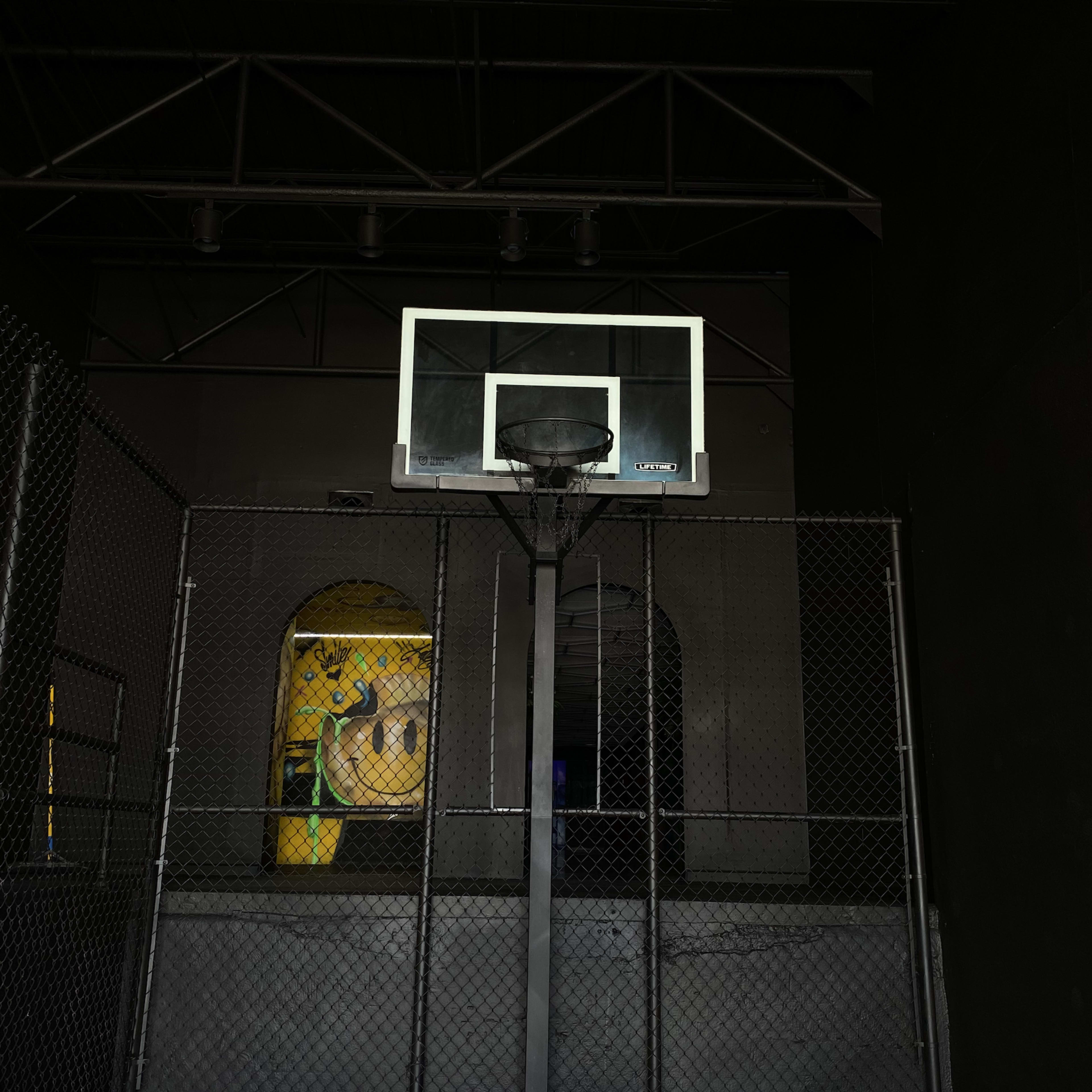 A basketball hoop installed in a dimly lit indoor court enclosed by a chain-link fence, with a large mural partially visible in the background.