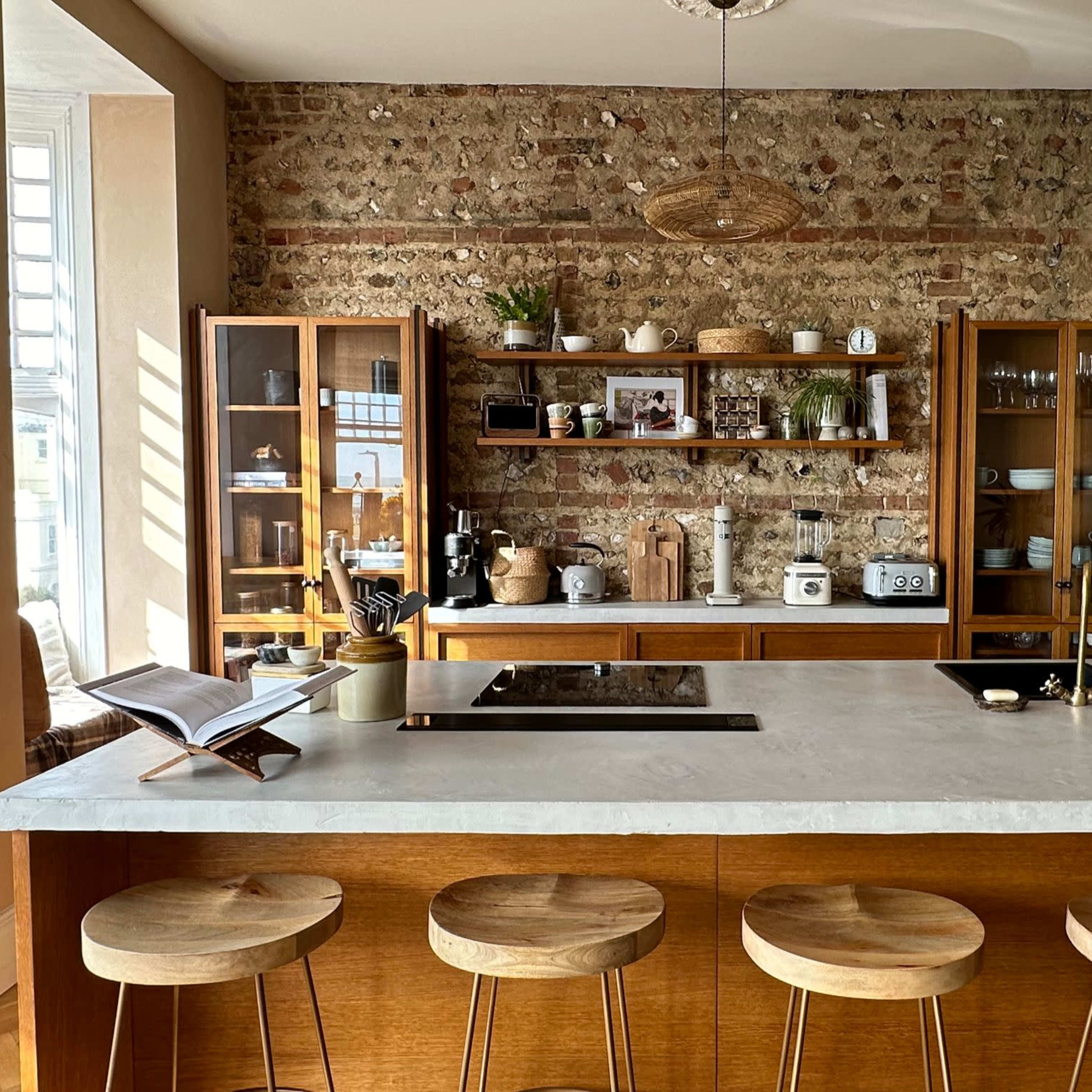 A modern kitchen features a marble countertop island with four wooden stools, surrounded by open shelving displaying various kitchen items against a textured brick wall.