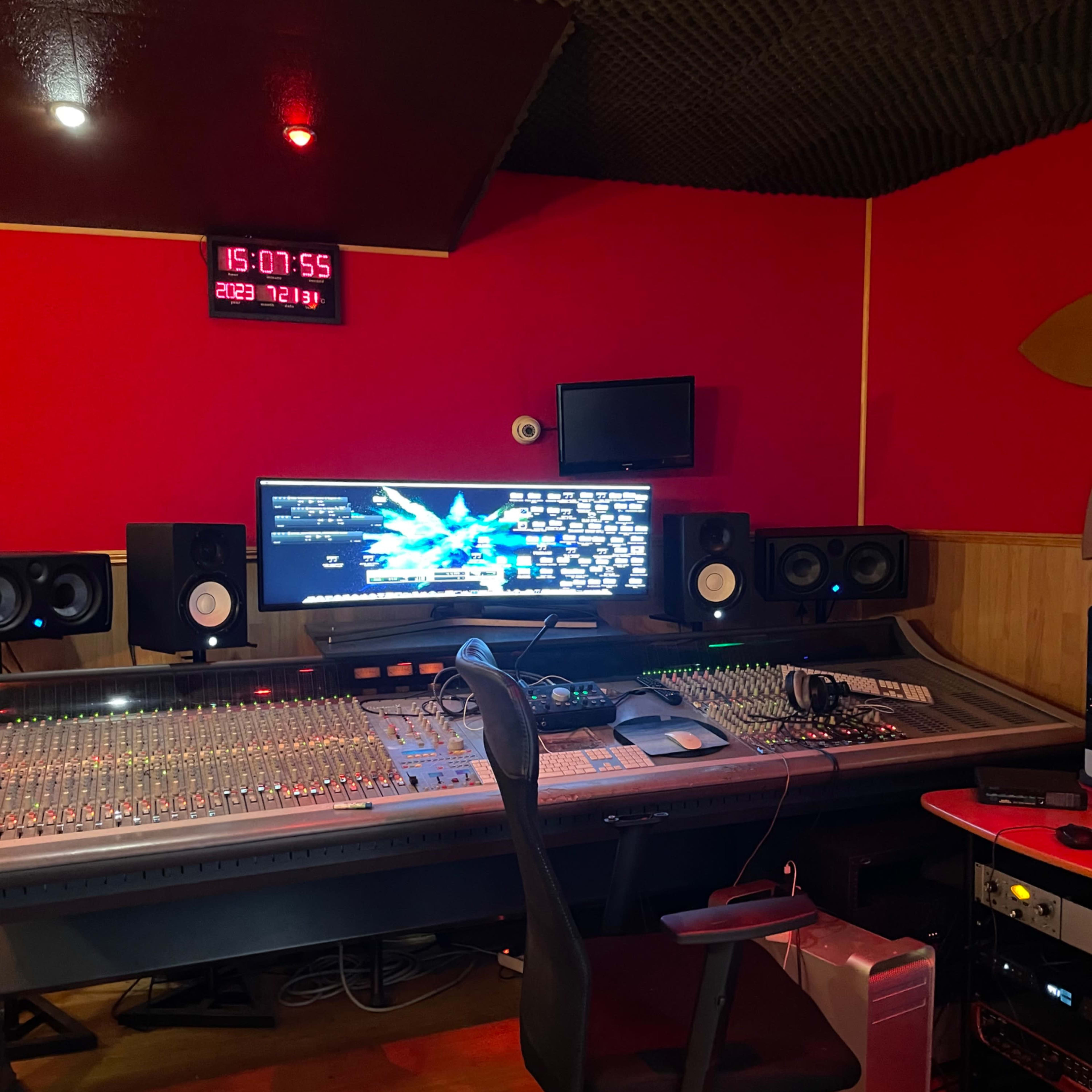 A music studio featuring a large mixing console, computer monitor, and sound equipment against a red wall and acoustic paneling.