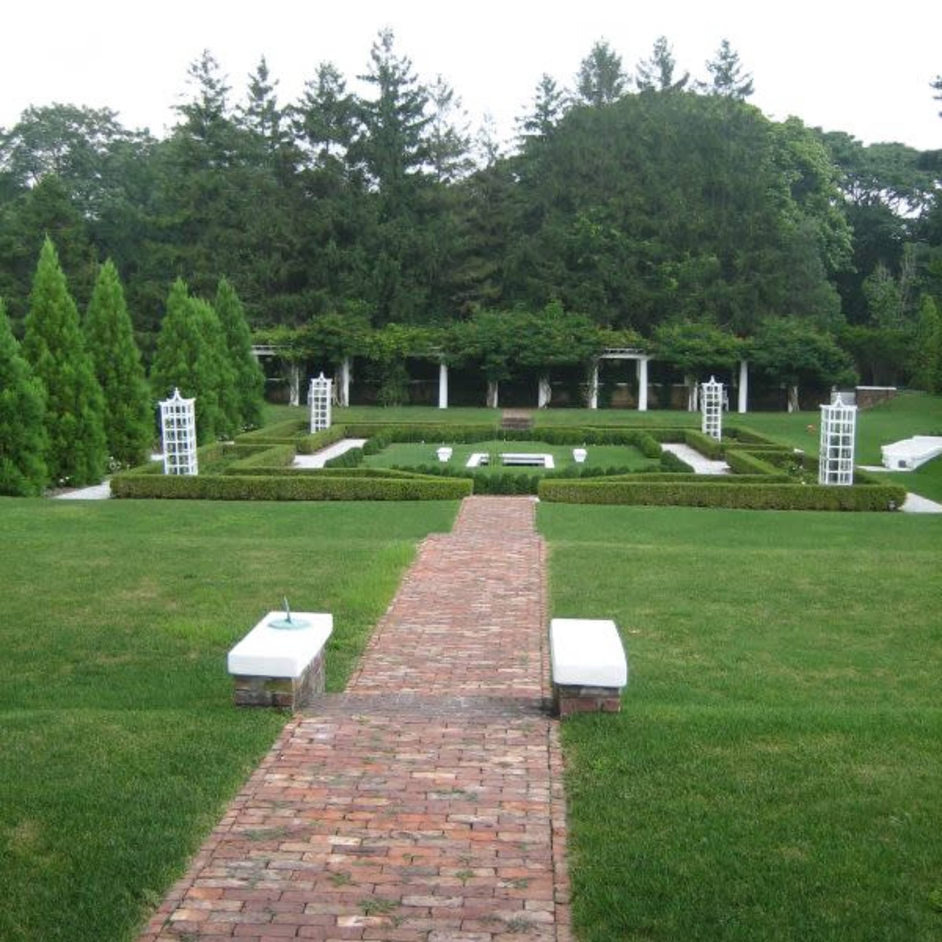 A landscaped garden with neatly trimmed hedges, a brick pathway leading to the center, and white structures positioned along the edges.