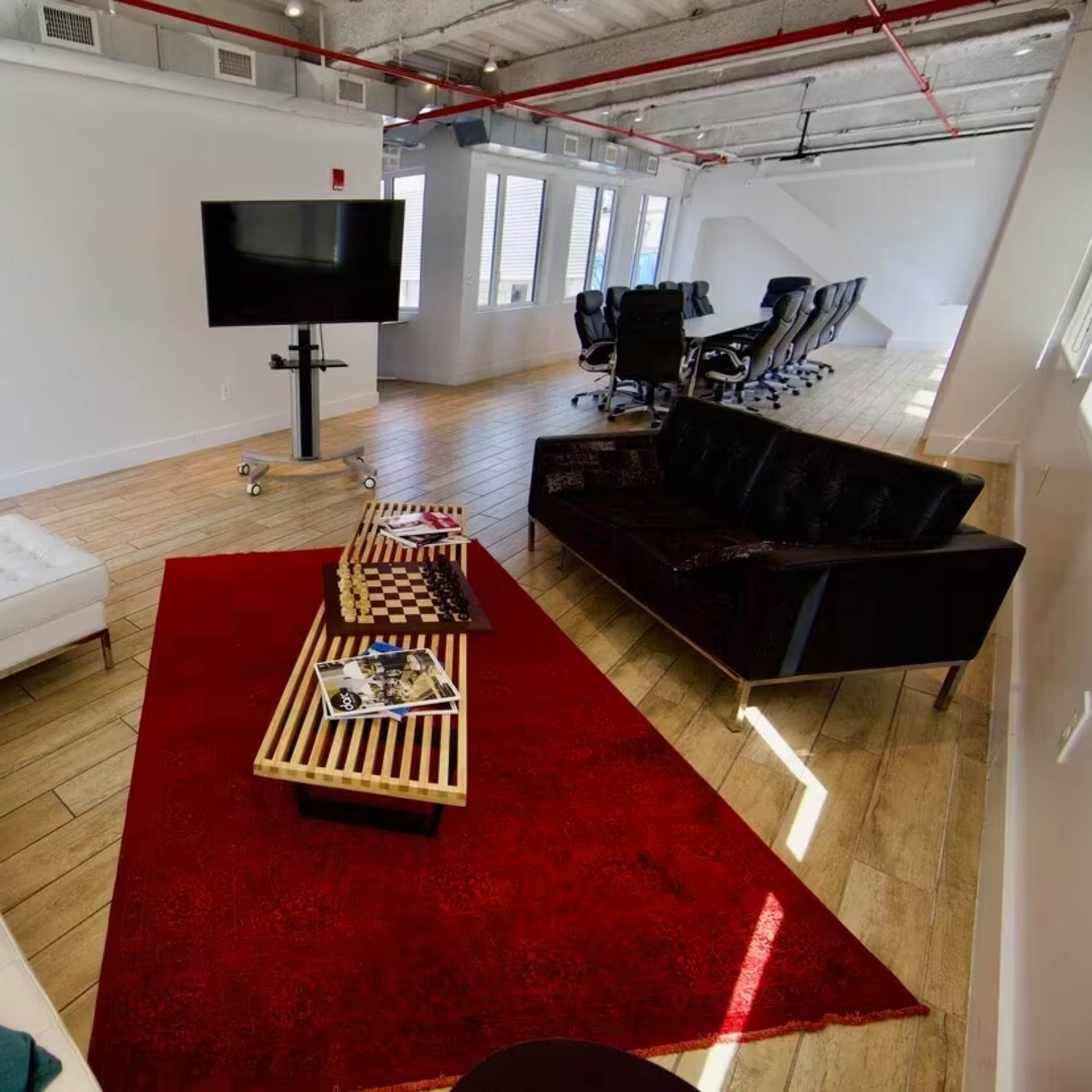 A modern office lounge area featuring a white sofa, a black couch, a red area rug, and a television mounted on a stand.
