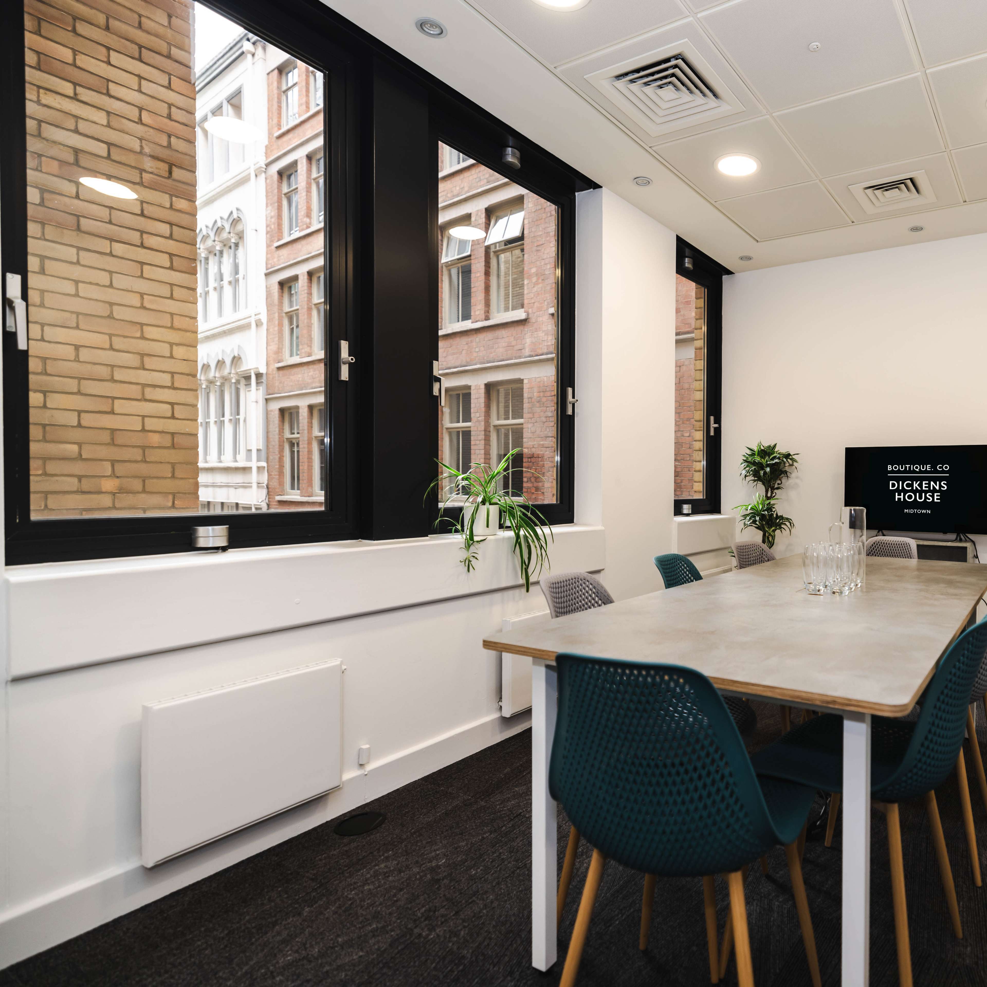 A modern meeting room with a long table surrounded by chairs, large windows providing natural light, and a television mounted on the wall.