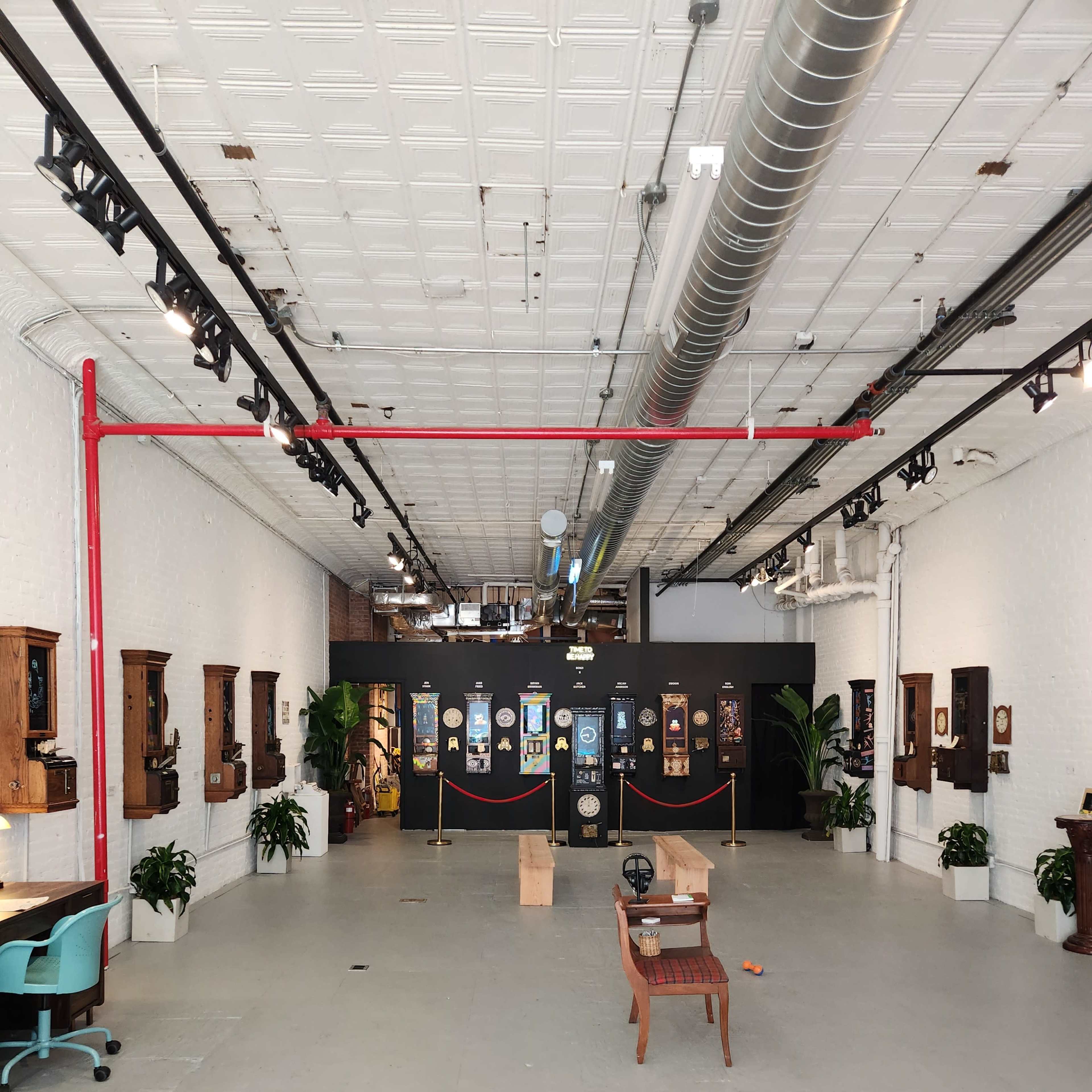A spacious gallery with white walls, high ceilings, and various framed artworks displayed against a black backdrop, along with potted plants and a simple wooden table in the center.