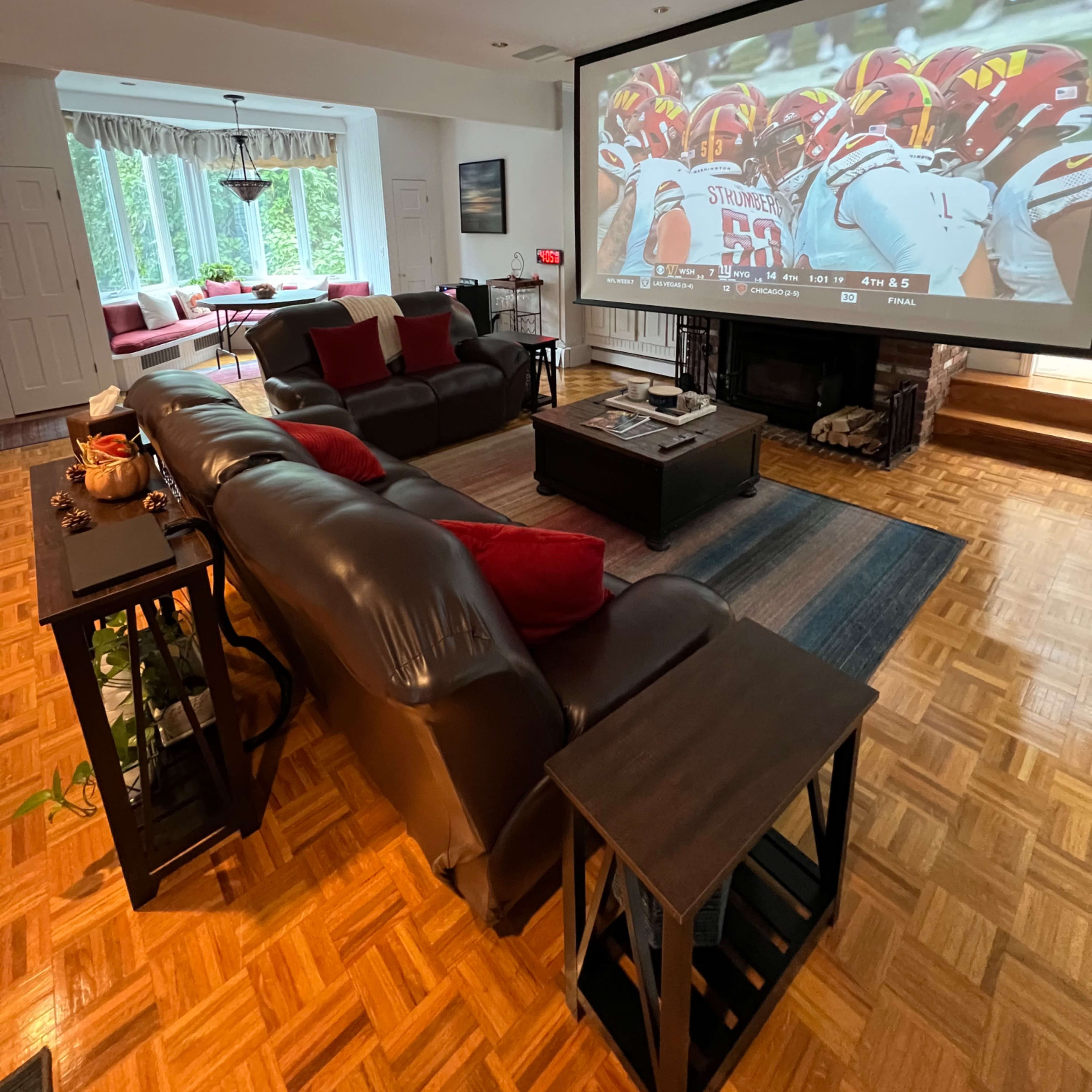 The living room features a large projector screen displaying a sports game, with dark leather sofas and wooden coffee tables arranged on a parquet floor.