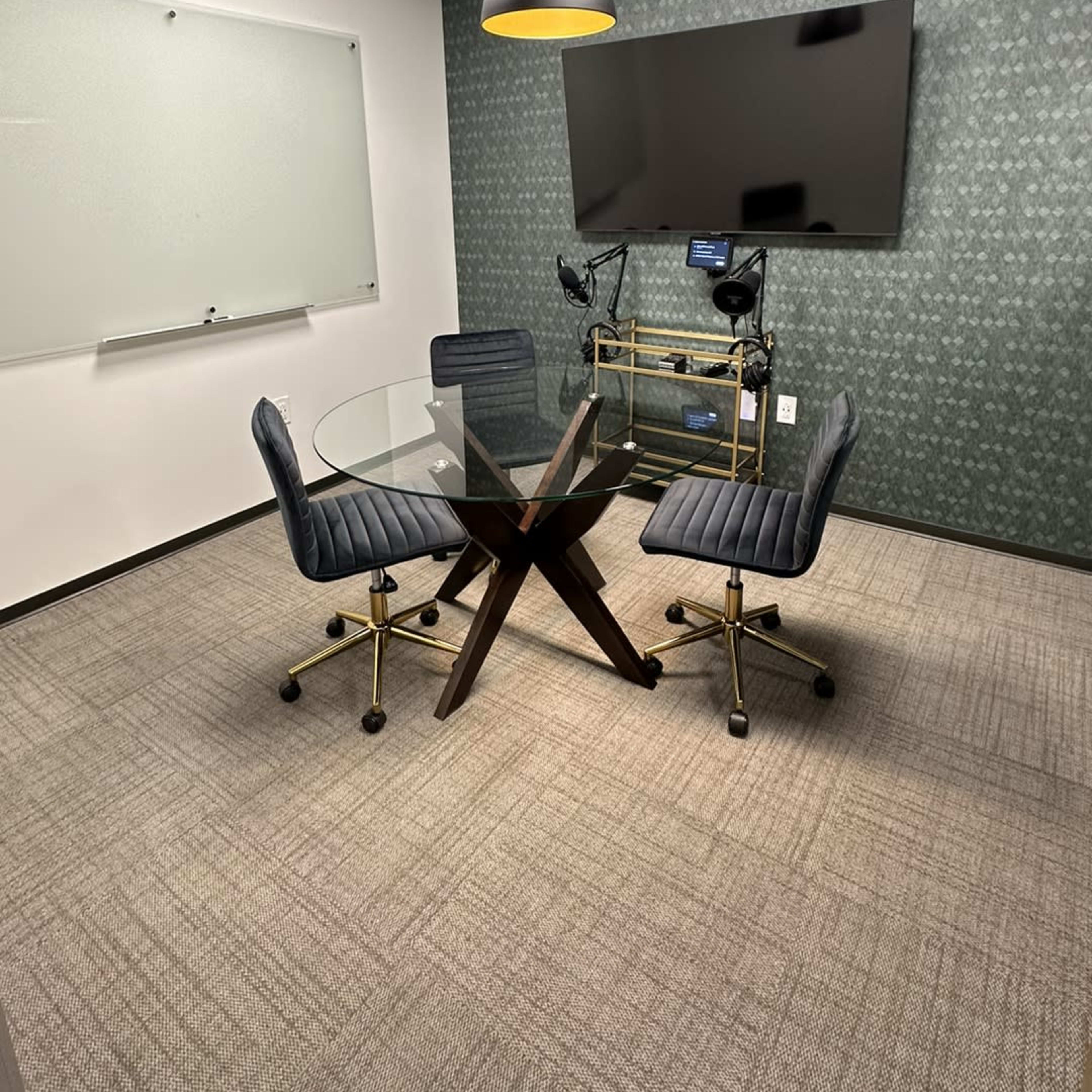 A small conference room featuring a glass table with four chairs, a wall-mounted television, and a whiteboard.