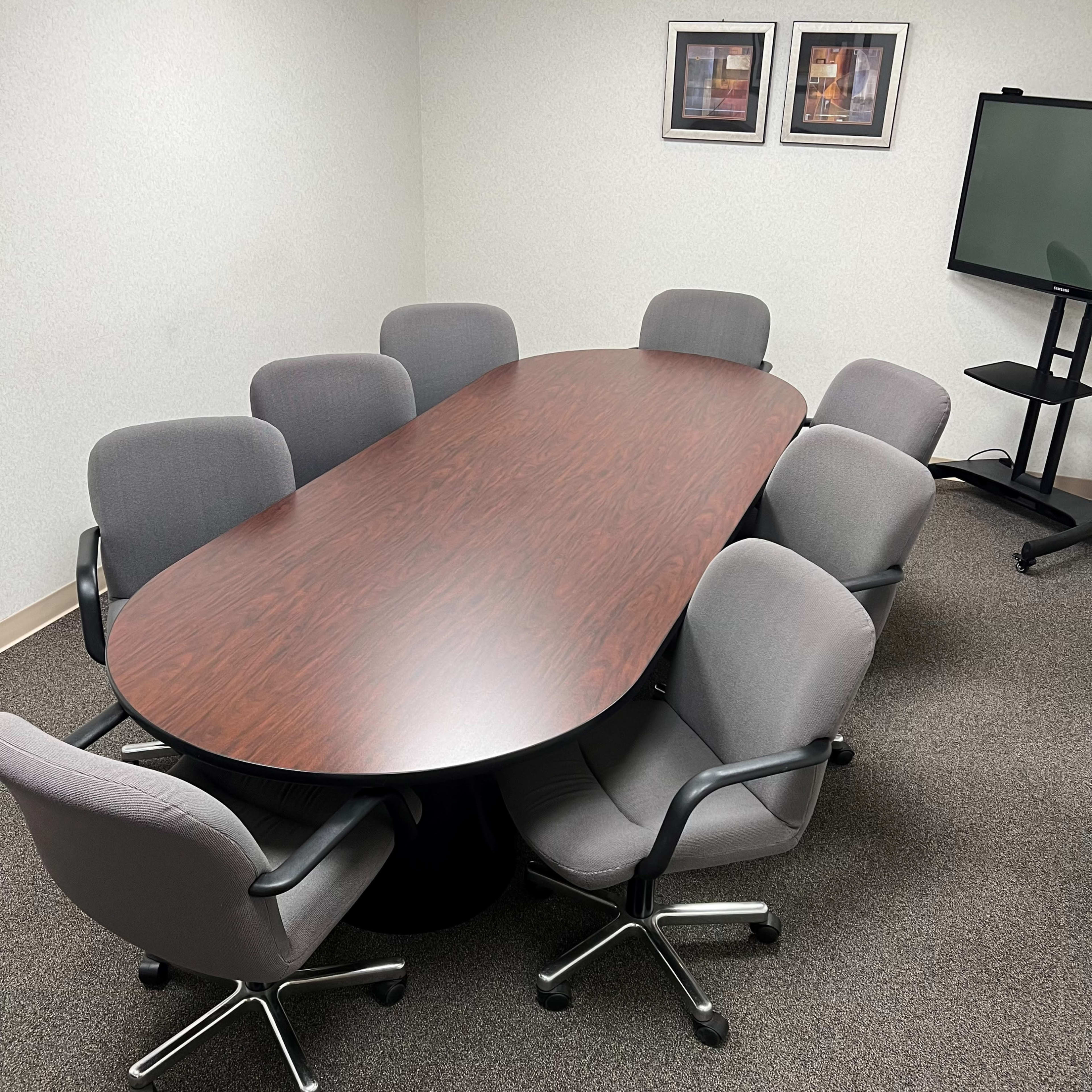 A large oval wooden conference table surrounded by eight gray office chairs is set up in a meeting room with a TV stand and framed pictures on the wall.