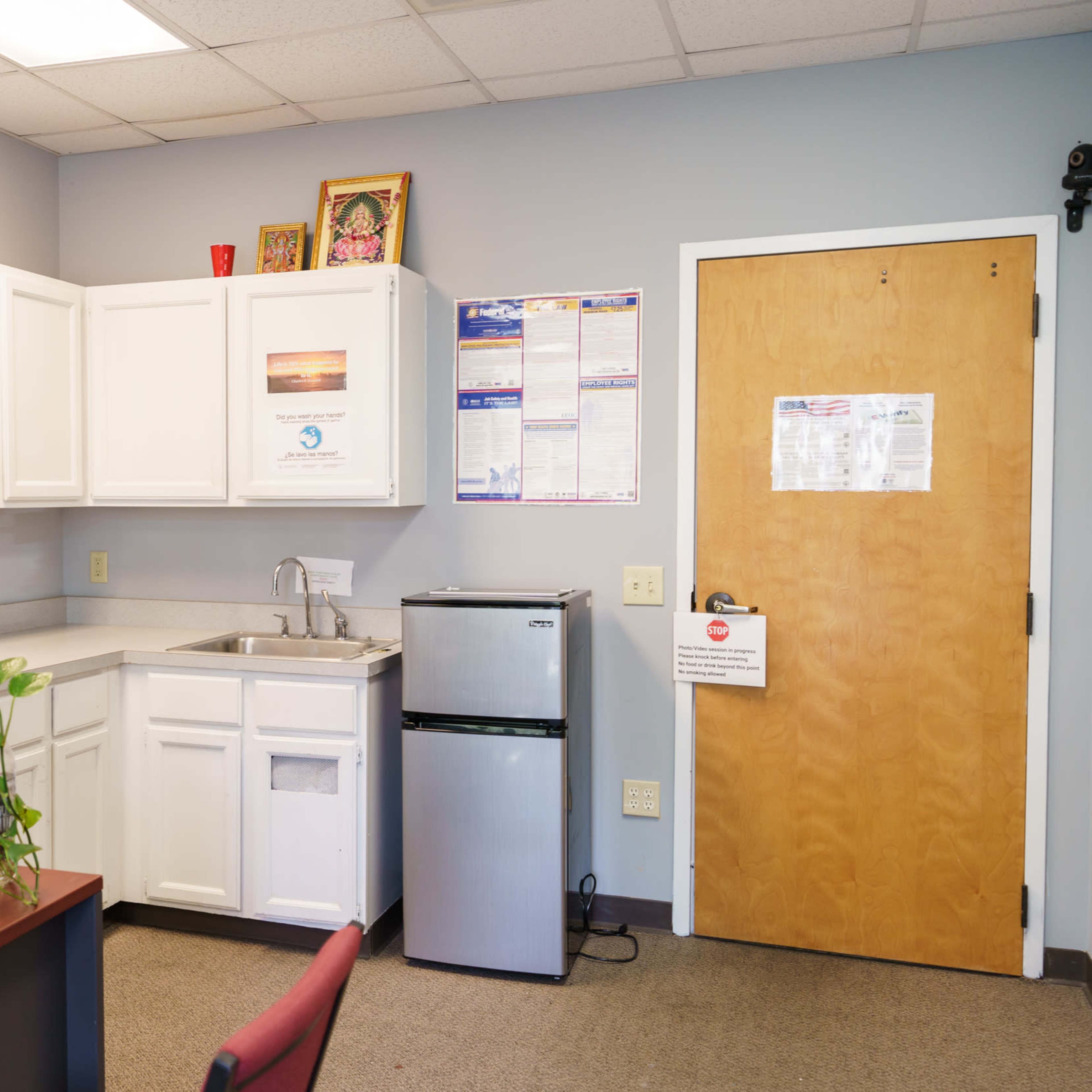A small office kitchenette featuring a sink, a mini-fridge, a desk with a computer, and a door with various safety notices.