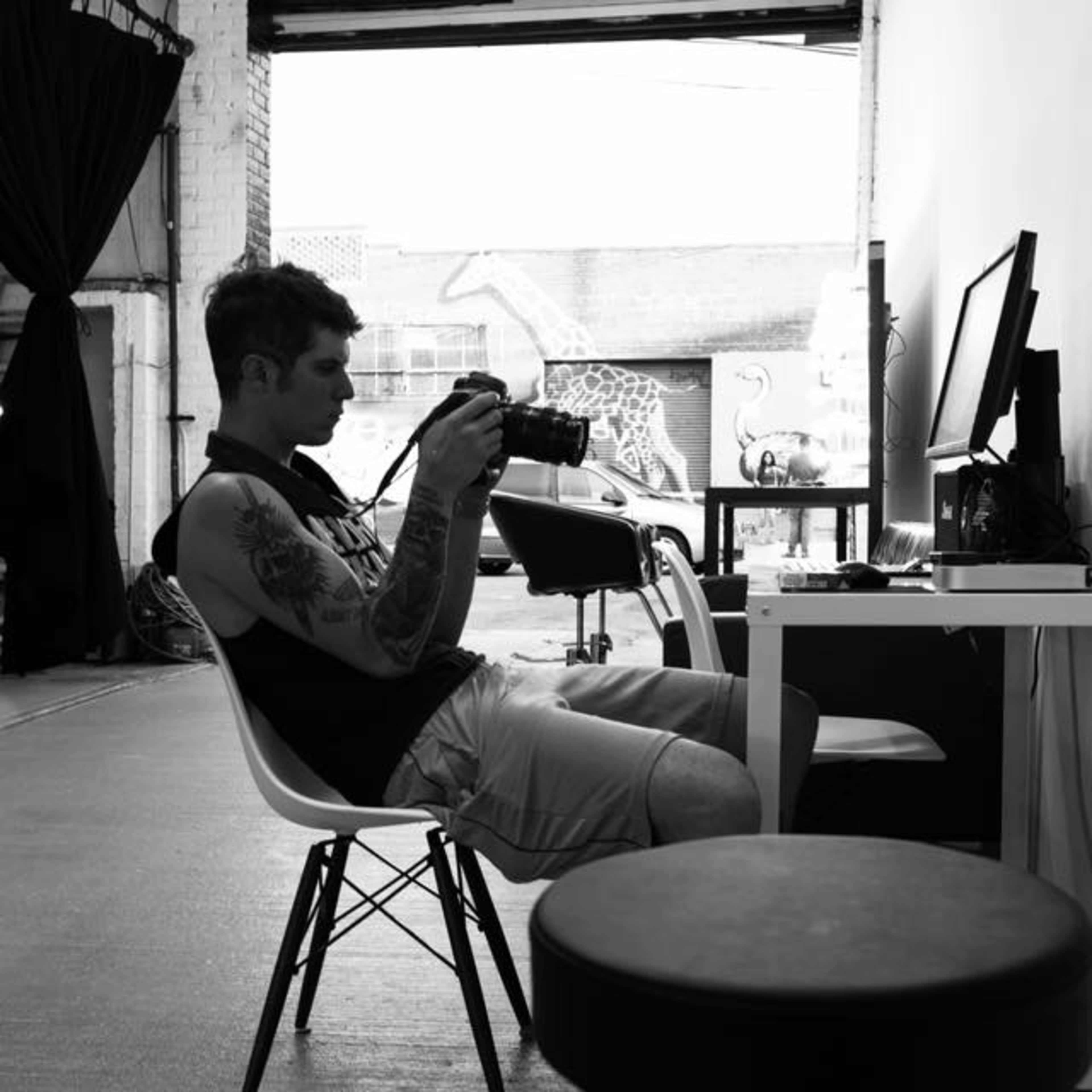 A person sitting on a chair in a well-lit room is examining a camera while a computer and a stool are nearby.