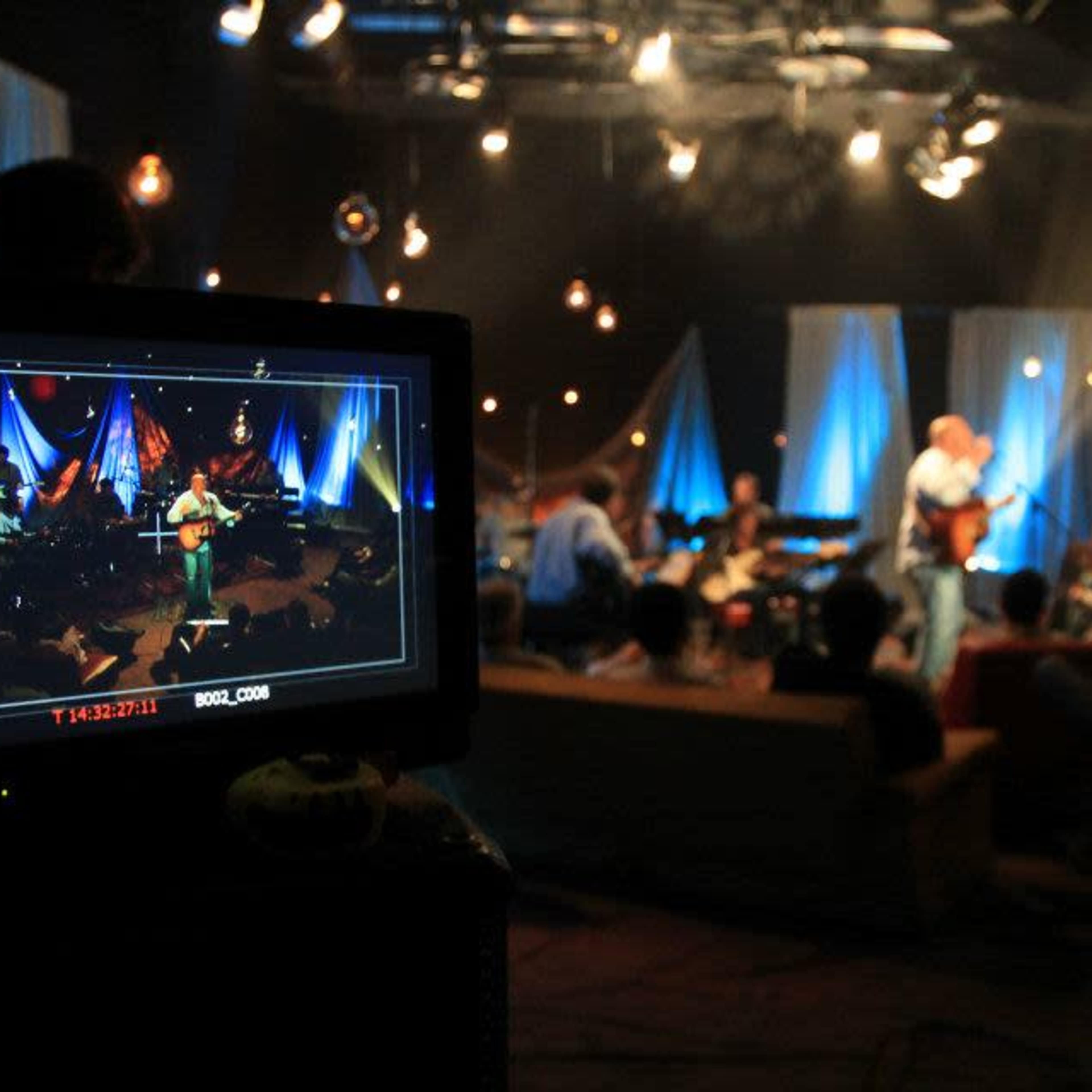 A musician performs on stage while an audience watches, as captured on a video monitor in the foreground.
