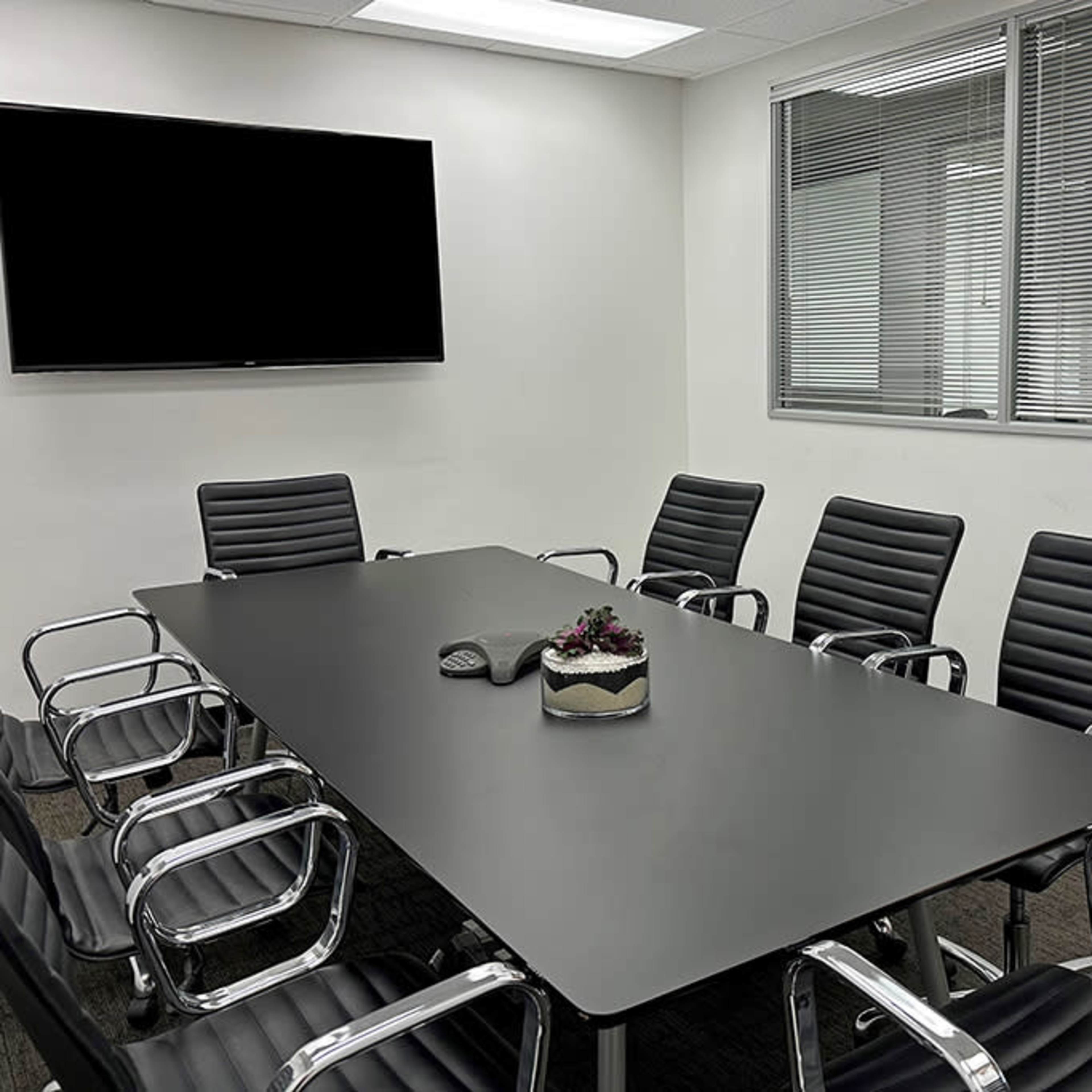 A modern conference room features a long black table surrounded by eight black leather chairs and a wall-mounted television.